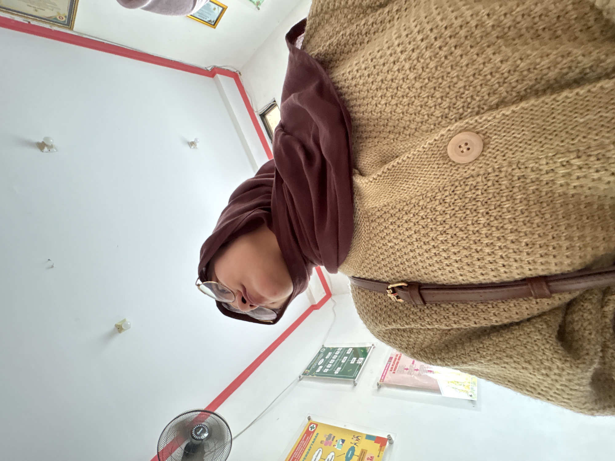 In a brightly lit room, a person wearing a brown hijab and a tan knitted cardigan is captured from a low angle, looking upwards. The person is wearing round-rimmed glasses and has a brown bag strap visible across their chest. The environment appears to be an indoor space with white walls adorned with various posters and a red-trimmed ceiling. A black oscillating fan is mounted on the wall to the left, and a light fixture is visible on the ceiling. The scene suggests a public facility, possibly a health clinic or office, given the informational posters on the wall. The lighting is bright, casting subtle shadows and highlighting the textures of the clothing and the room's surfaces. The overall impression is of a candid moment, perhaps taken while waiting or observing something above. The location is identified as Kebonsari, Indonesia.