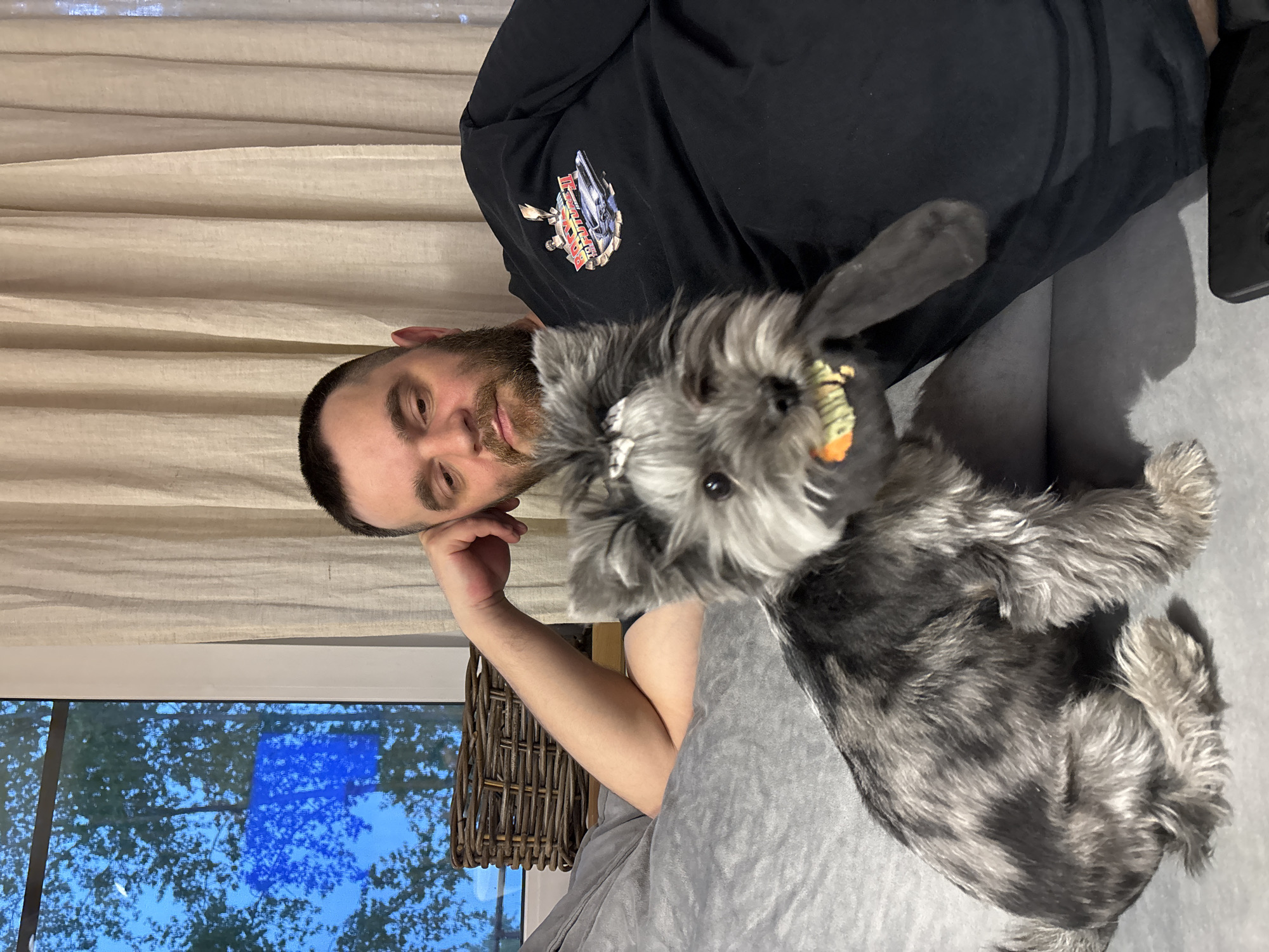 A man with a beard rests his head on his hand beside a gray schnauzer-like dog with a hair bow. The man is wearing a black T-shirt with a graphic. They appear to be indoors on a gray couch. In the background, there is a window showing trees and a bright blue sky. Next to the window is a wicker basket. There is a curtain hanging behind the man. It is impossible to verify the location in the city of Chaikovsky, Russia, from the given image.