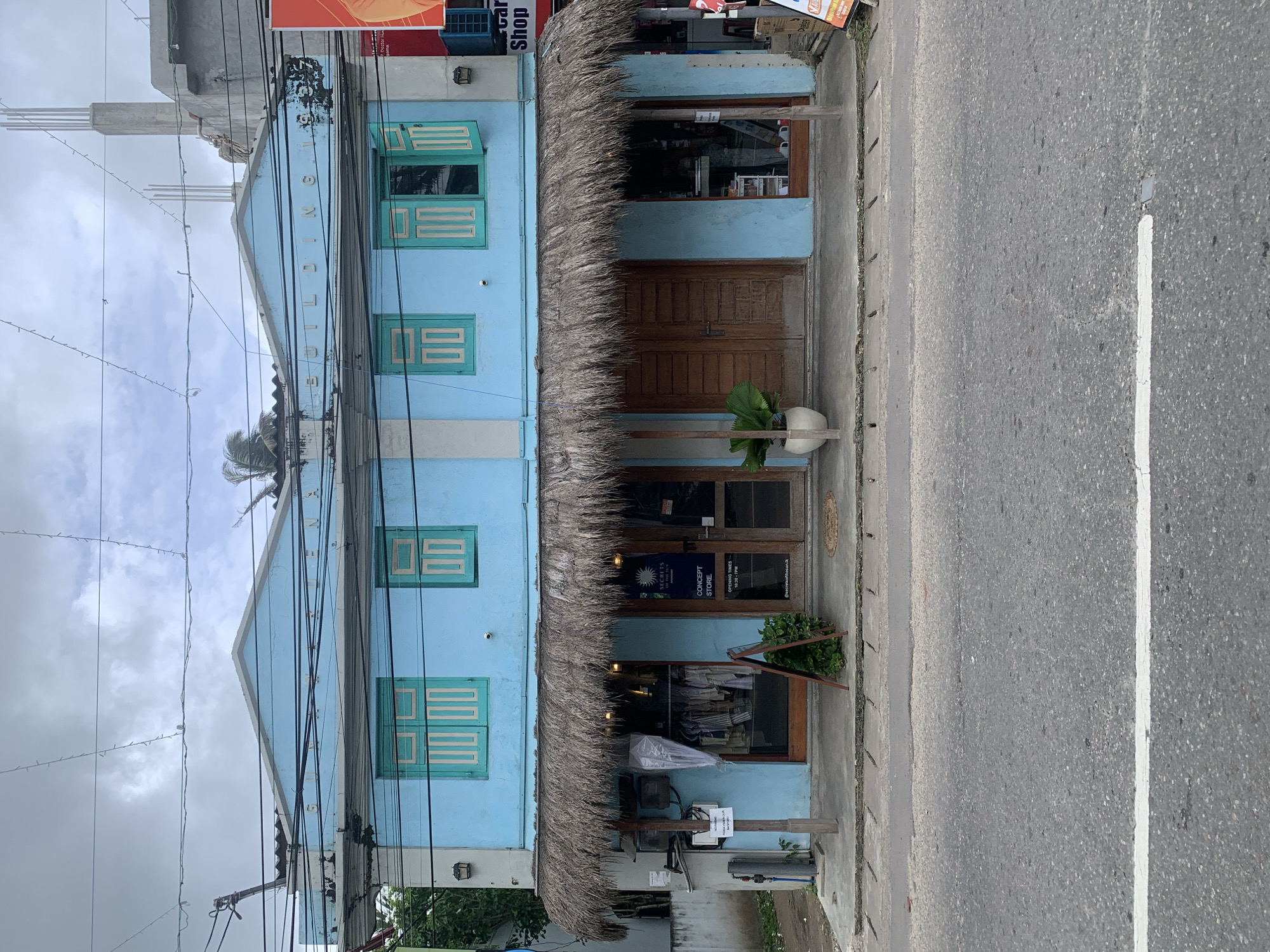 The image depicts a street view of a light blue two-story building in Ahangama, Sri Lanka. The building has a thatched roof awning over its storefront. The storefront has two entrances and two display windows. One display window reveals clothing racks with apparel, while the other shows various items. A plant in a white pot is situated to the left of the right entrance. The building's upper floor features two windows with teal shutters that are partially open. A sign on the building reads "NA BUILDING". The street in front of the building is paved with asphalt and has a white stripe. The sky above is overcast with visible power lines crisscrossing the scene.