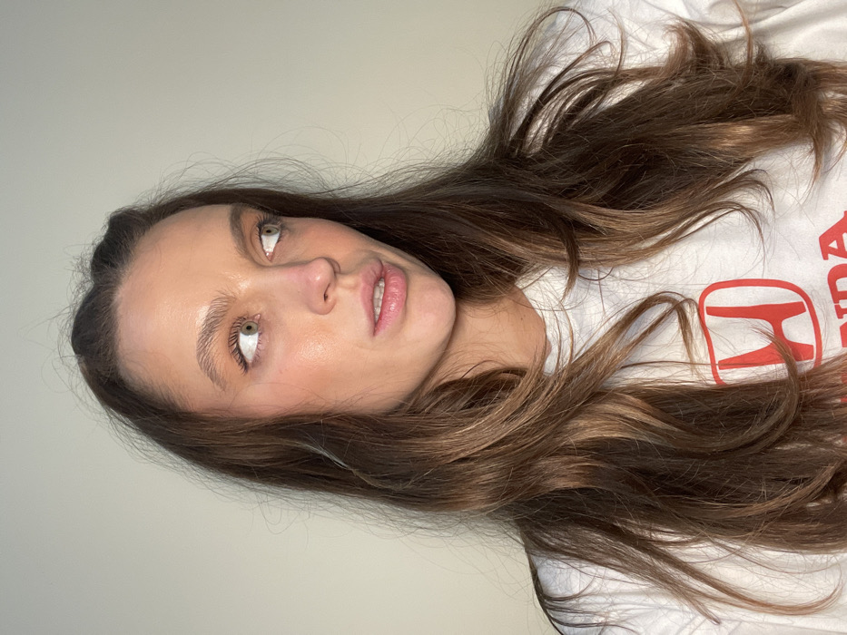 A woman with long brown hair and fair skin looks up and to the left. She's wearing a white T-shirt with a red Honda logo partially visible. Her expression is neutral, with a slight hint of a smile. Light makeup highlights her eyes and cheekbones. The background is a plain, light-colored wall. The image has a slightly rotated orientation.