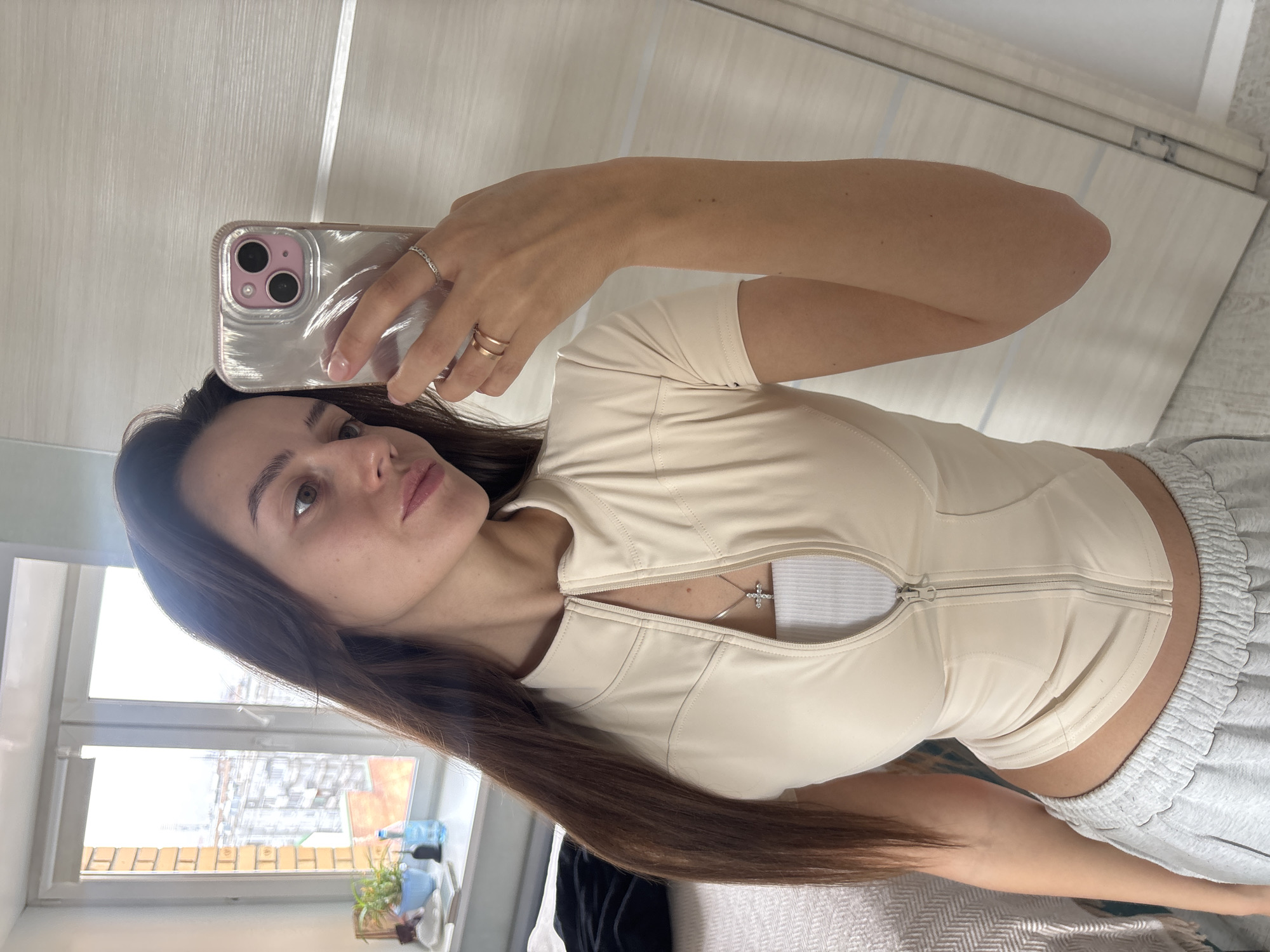 A woman with long, brown hair is taking a selfie. She is holding a smartphone in her right hand, which has a silver case with pink accents around the camera lenses. She has two rings on her fingers. The woman is wearing a fitted, beige-colored zip-up shirt and gray sweatpants. The zip-up shirt is partially unzipped. A silver cross necklace is visible beneath the shirt. She is standing in what appears to be a room with white walls and a light-colored door. A window is visible in the background, with a view of a city. Based on the buildings in the background, this is possibly a location in the city of Krasnoyarsk, Russia. There are plants and other small objects on the windowsill. The photo is taken at a slight angle, and the lighting suggests it is daytime.