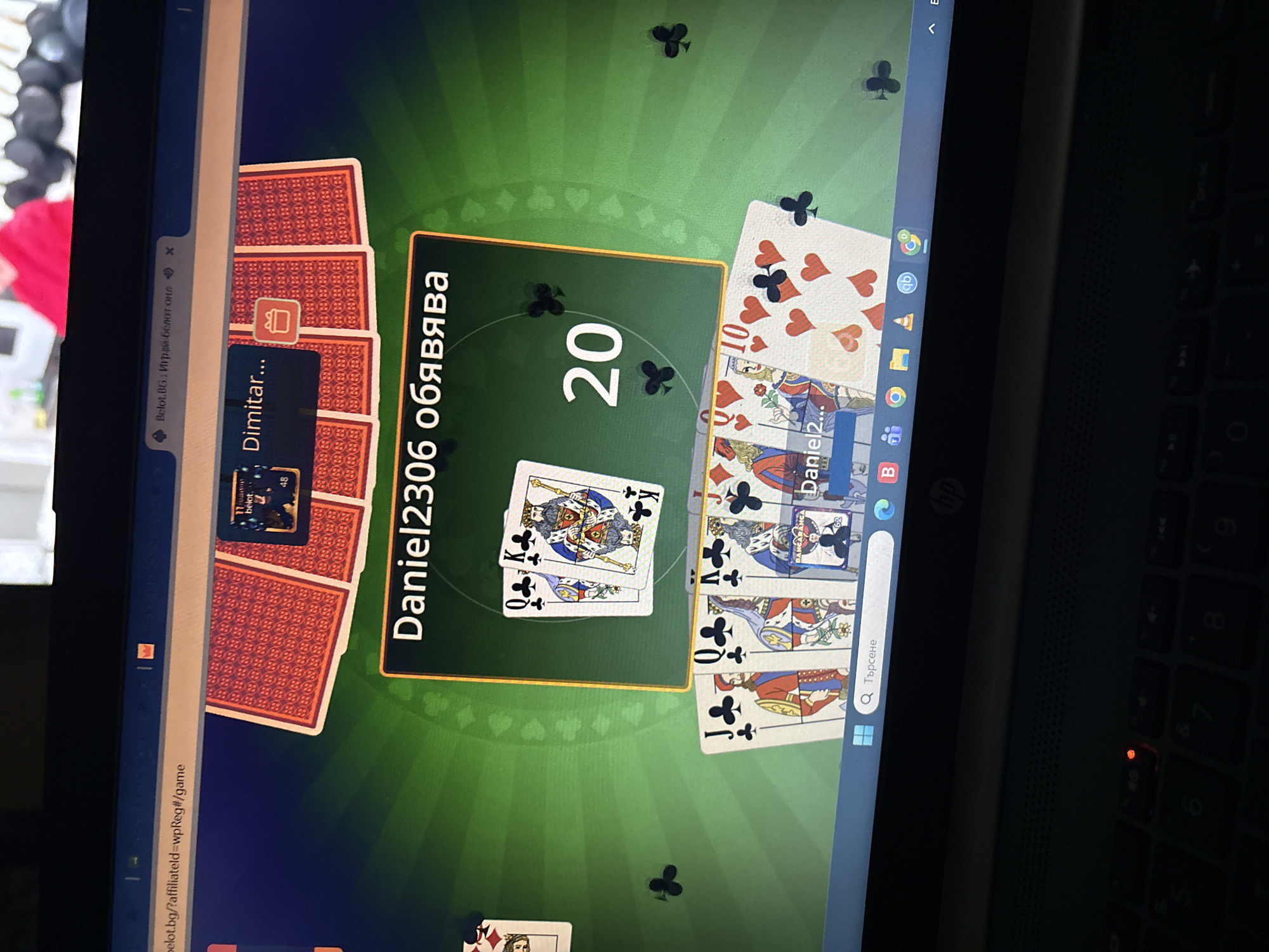 The image shows a laptop screen displaying an online card game. The game appears to be Belot, a popular card game in Bulgaria. The screen shows two players: "Dimitar..." and "Daniel2306". A card pile is visible on the left side of the screen. In the center of the screen, “Daniel2306” has 20 points. Cards are visible on the table. The user is located in Sofia, Bulgaria, as the language on the screen is Bulgarian, and the website domain shown is ".bg", the domain extension for Bulgaria.