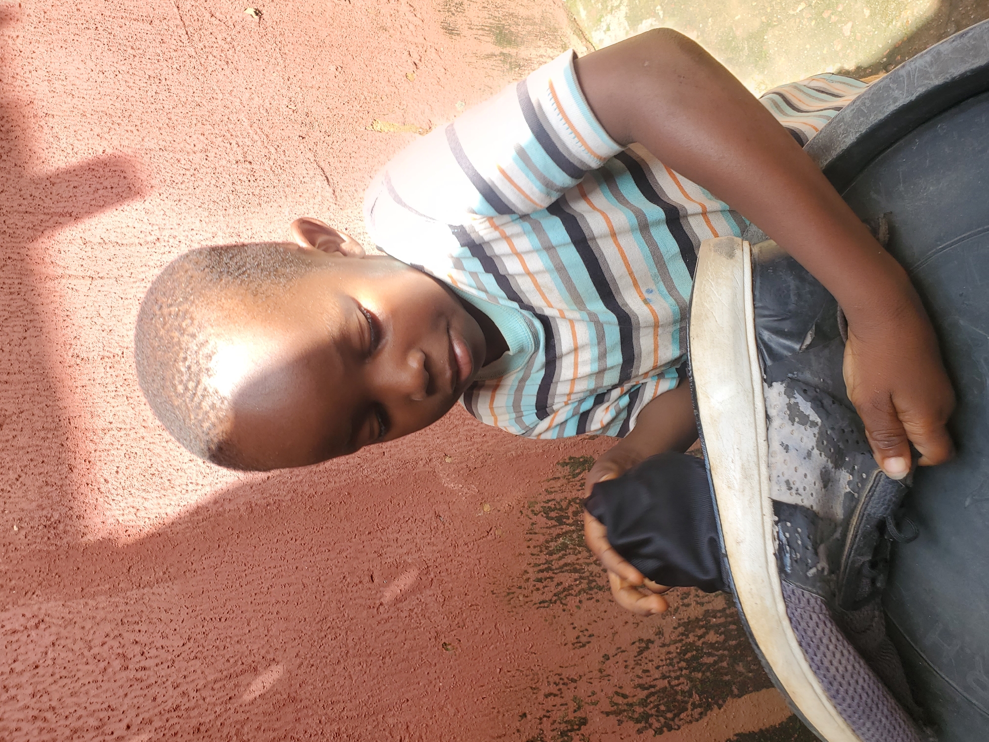 A young boy with dark skin and a short haircut is seen from the chest up, looking down at a pair of black shoes. He is wearing a horizontally striped t-shirt with colors of white, light blue, orange, and black. His right hand is holding a black cloth object near the shoes, while his left hand is touching the sole of one of the shoes. The shoes appear to be worn and are placed on a black circular object, possibly a container. The background is a textured, reddish-brown wall with some patches of green algae or moss and sunlight casting shadows. The ground beneath the boy is visible and appears to be a light-colored, uneven surface. The scene is captured outdoors, likely in a residential area of Jos, Nigeria, given the architectural style of the wall and the general environment.