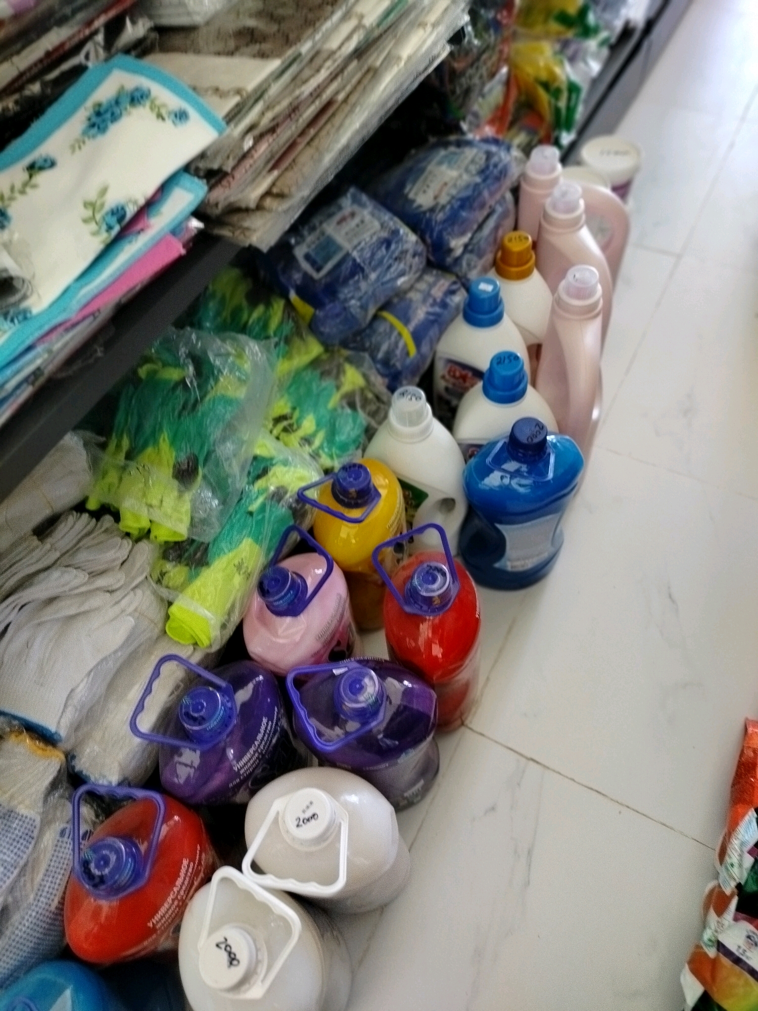 The image depicts a retail setting, likely a small store or market stall, displaying a variety of household cleaning products and miscellaneous items. The products are arranged on shelves and on the floor. Prominently featured are numerous plastic bottles of cleaning liquids in various colors, including blue, red, yellow, and purple. Some bottles have handles, and others are larger jugs. There are also stacks of white cloth gloves, and a few packaged items with blue and green hues, possibly cleaning cloths or sponges. In the background, more products are visible, including what appear to be fabric items like towels or sheets with floral patterns. The flooring is light-colored tile. The overall impression is one of a well-stocked, albeit somewhat cluttered, retail space. The scene is devoid of any people. The specific location of Көлкент, Kazakhstan, is not discernible from the visual information in the image itself.