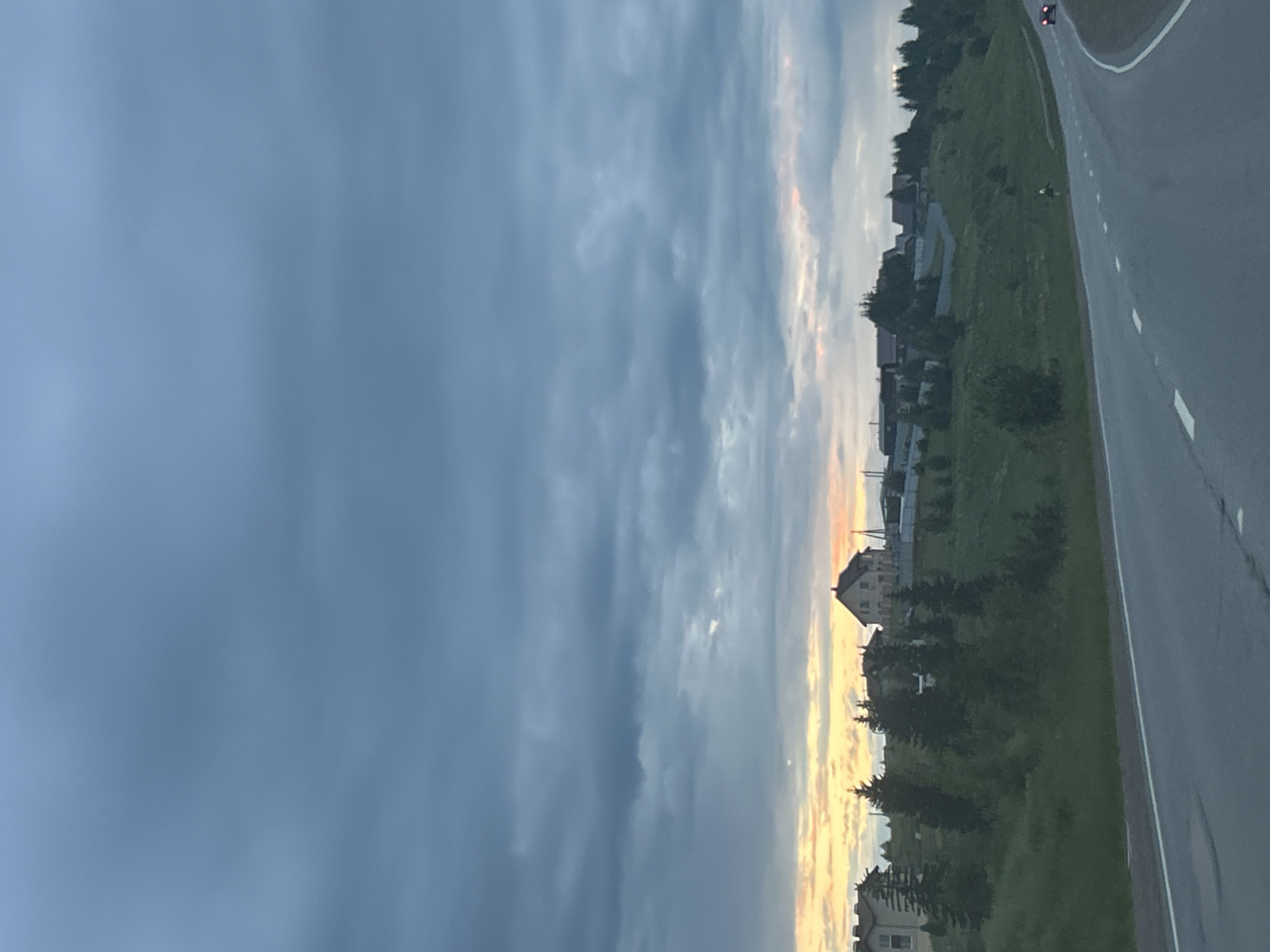 The image depicts a rural road scene near the village of Staroye Martyanovo, Russia, under a cloudy sky. A paved road curves into the distance on the right side of the frame, with white dashed lines marking the lanes. To the left of the road, a grassy hillside is dotted with trees and several houses with red roofs. The sky is predominantly a muted blue-gray with streaks of orange and yellow near the horizon, suggesting either sunrise or sunset. There are no people visible in the image, but two vehicles, one red and one white, are present on the road. The overall atmosphere is calm and serene.