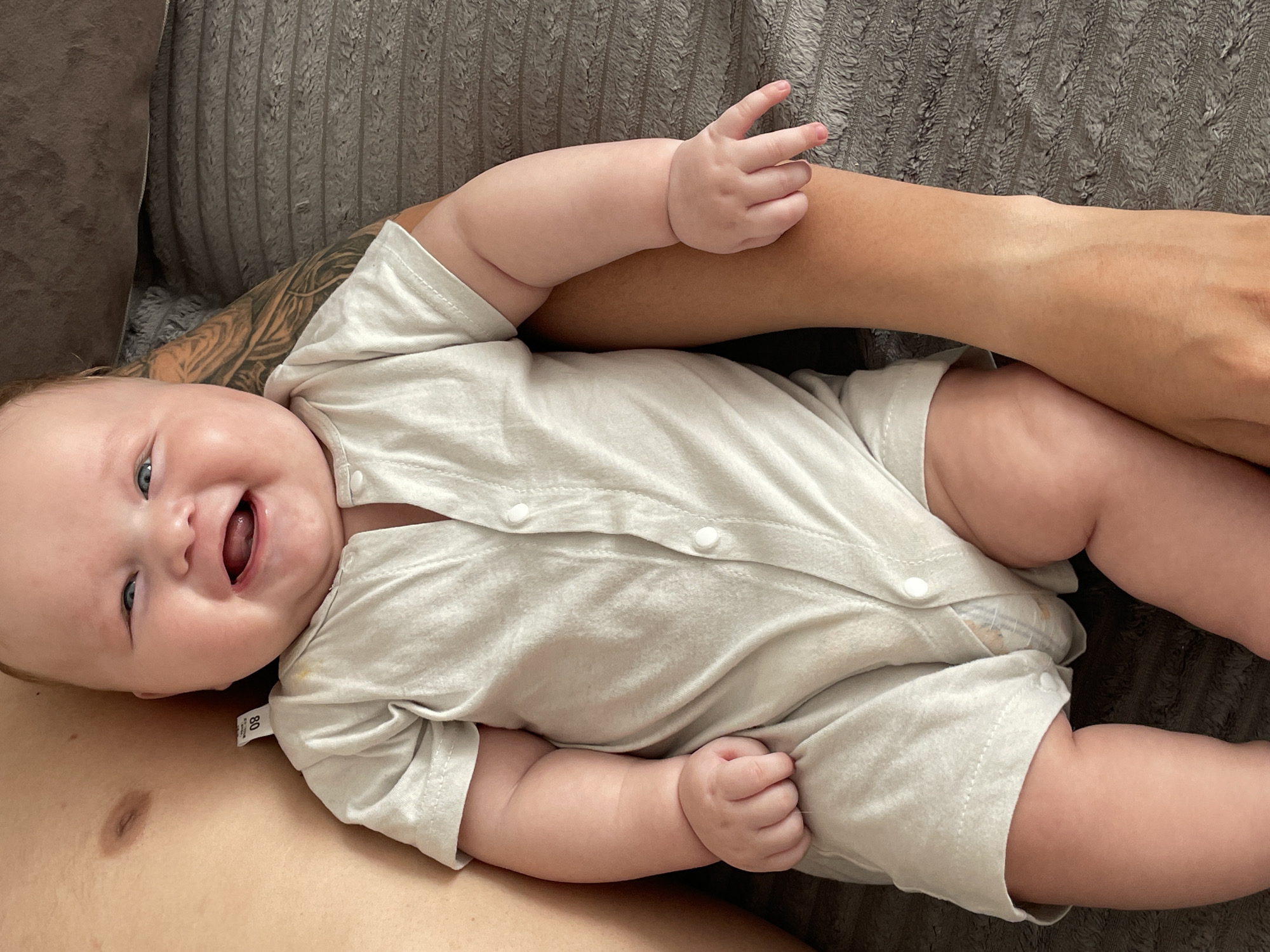 A baby is lying on a textured, gray surface, possibly a couch or blanket. The baby is wearing a light beige romper with white buttons. The baby has light skin, blue eyes, and a wide, happy smile with its mouth open and tongue visible. Visible on the baby's romper is a tag with the number "80" printed on it.
An adult's arm, with visible tattoos on the forearm, is positioned behind the baby's head. Another adult's arm is resting underneath the baby's body, partially obscured by the romper. The adult's skin tone appears to be lighter than the baby's. The overall lighting suggests it is daytime, and the setting appears to be indoors. The mood conveyed is one of happiness and comfort, with the baby's expression being particularly joyful. There are no specific weather cues or strong location indicators beyond the potential indoor setting.