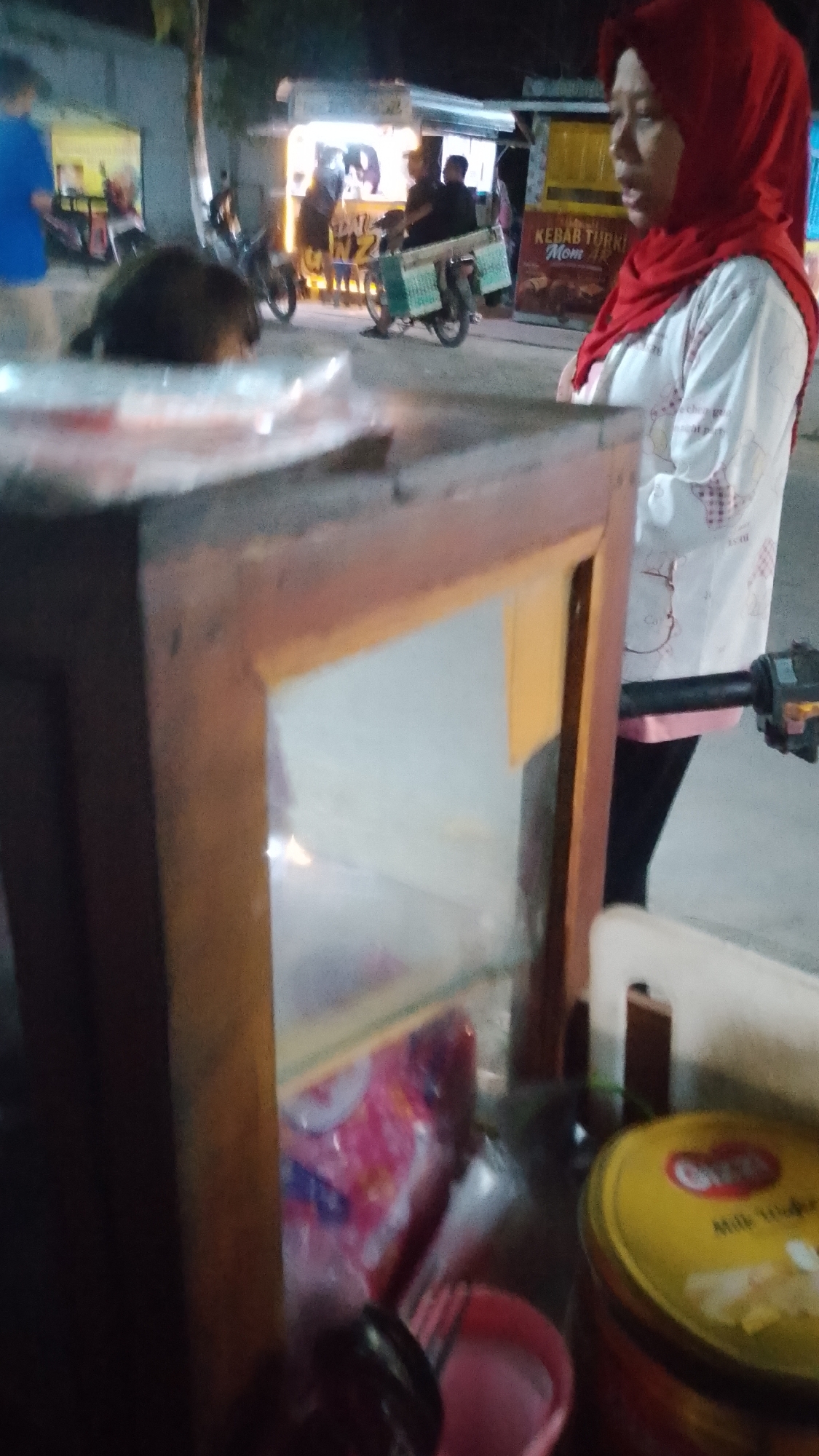 An outdoor scene at night in Karanglangu, Indonesia, shows a food stall with bright lights and several people. In the foreground, a woman wearing a red hijab and a white shirt with a pattern is standing, looking to her right. Behind her, a wooden display case with glass shelves holds various items, including pink packaged goods and what appears to be a yellow tin can with text. To the left of the display case, a dark, furry object is visible, possibly an animal. In the background, a food stall is illuminated, with people visible behind the counter. A motorcycle with green crates attached to its rear is parked nearby. Another person in a blue shirt is standing to the far left. The overall environment suggests a busy street food area.