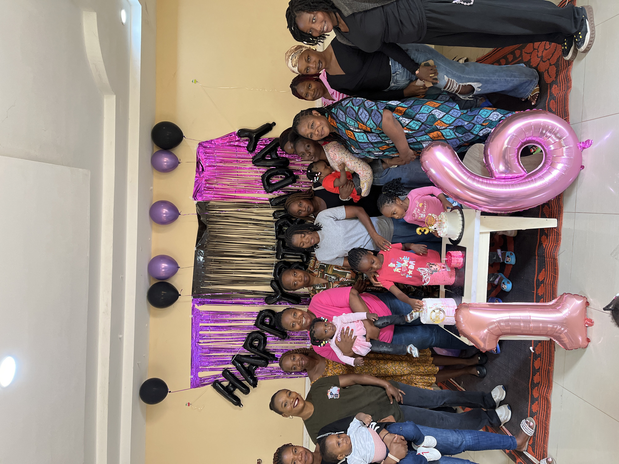 This image depicts a celebratory gathering of individuals within an indoor room in Kuru Karama, Nigeria. The scene features a group of 16 adult women and approximately 6 young children, ranging from infants to toddlers. The adults, primarily of dark complexion, are dressed in a mix of casual attire including t-shirts, dresses, patterned garments, and jeans, with various hairstyles including natural styles, braids, and headscarves; they are positioned for a photograph, with several women holding children. Birthday decorations are prominently displayed, including black foil balloons spelling "HAPPY BIRTHDAY" against a backdrop of purple and black shimmering metallic foil curtains, and additional black and purple round balloons floating near the ceiling. In the foreground, two large pink metallic foil balloons shaped like the numbers '1' and '6' rest on a patterned rug, indicating a 16th birthday celebration. A small white rectangular table in the center holds at least three birthday cakes, including one with a "3" topper and another with a gold topper, alongside a pink gift box. The floor consists of light-colored tiles partially covered by a dark-colored rug with an orange pattern, while the walls are painted a plain, light yellowish-beige. The white ceiling features visible seams and recessed lighting fixtures, illuminating the scene. A young girl in a pink shirt stands by the table, looking at the cakes.