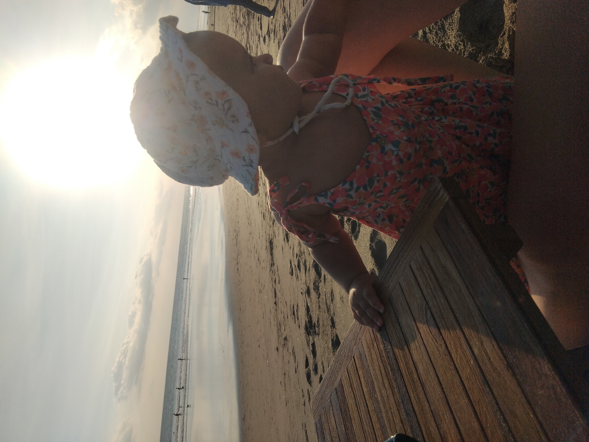 The image captures a peaceful beach scene in North Kuta, Indonesia, likely during sunrise or sunset, as indicated by the intense, low sun on the left side of the frame. The primary subject is a young baby or toddler, viewed from the side, wearing a white sun hat with a delicate floral pattern and a red/orange floral dress. The baby's head is turned towards the bright horizon, as if looking at the sun or the vast ocean.
The baby is positioned close to a wooden slatted surface, possibly a low table or part of a beach chair, with one hand resting on it. A partial view of an adult's limb (likely an arm or leg) is visible next to the baby, suggesting a parent or guardian is present.
The background reveals a wide, sandy beach stretching into the distance, meeting the calm ocean under a partly cloudy sky. Several small, silhouetted figures can be seen walking or standing along the distant shoreline, adding a sense of scale and life to the tranquil landscape. The overall lighting is warm and bright, characteristic of golden hour, creating a soft glow over the scene. There is no visible text in the image.
