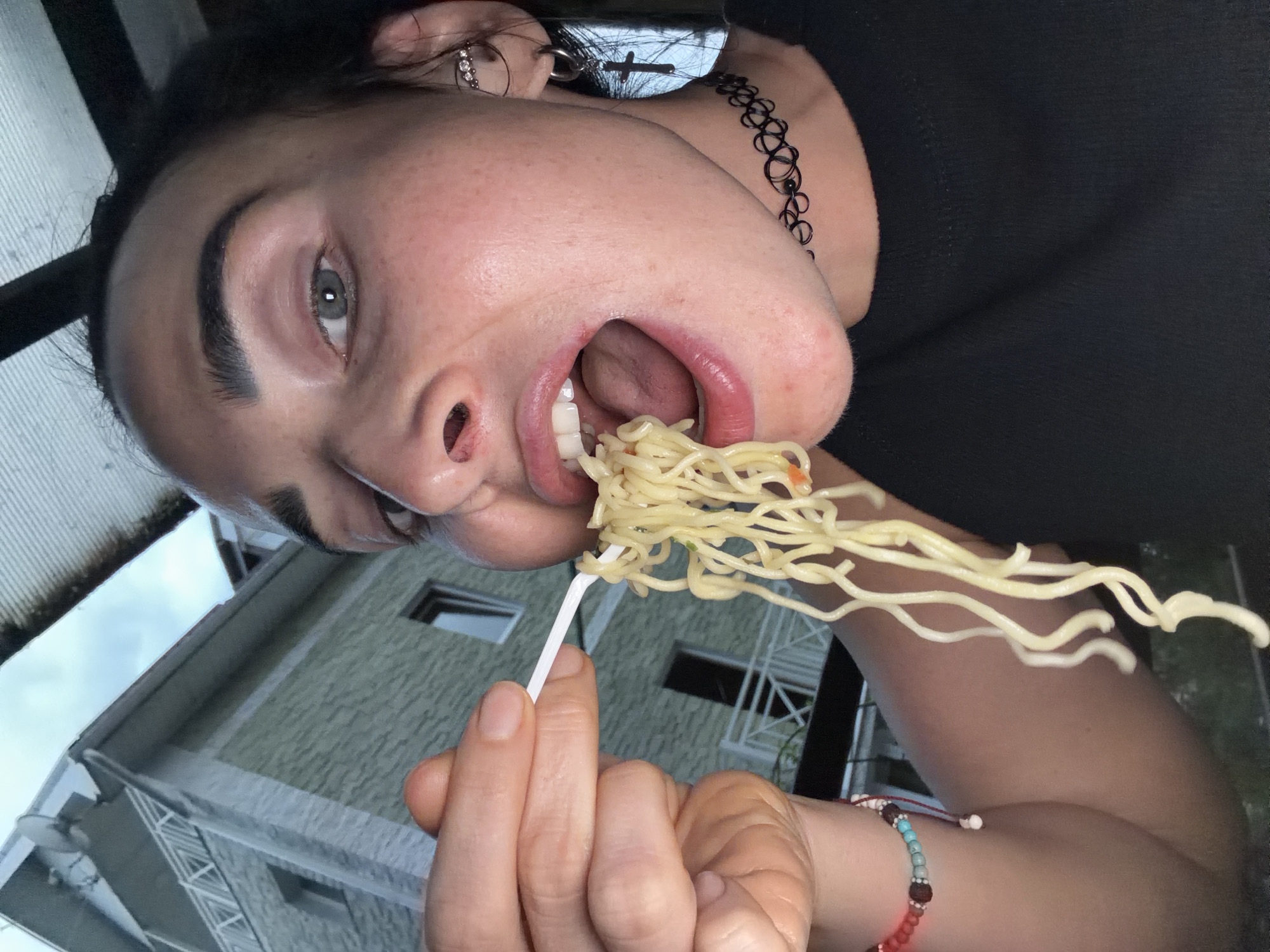 A close-up, low-angle shot captures a woman with her mouth open, eating ramen noodles with a white plastic fork. She is wearing a black t-shirt, a black choker necklace with a circular design, and a bracelet made of colorful beads on her left wrist. She has a cross earring with a ring attached in her right ear and a row of small jewels in her upper ear. Her eyebrows are dark and well-defined. In the background, a light green building with a white railing on a balcony and a satellite dish on the roof is visible. The sky appears overcast. The overall scene suggests an outdoor setting. It is not possible to determine the specific location within Sochi, Russia from the provided image.