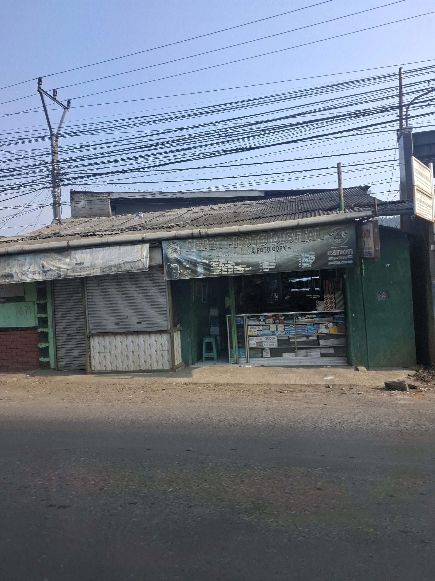 The image shows a street-level view of a commercial building in Karangtengah, Indonesia. The building appears to be a small business, possibly a photo studio and copy shop, given the signage visible. The facade is a mix of materials, including what looks like corrugated metal roofing and siding, some brickwork on the left, and tiled lower sections. There are closed metal shutters on a portion of the storefront. Inside the open section of the shop, shelves are visible, stocked with various items, including paper products. A green plastic stool is positioned outside the entrance. The street in front of the building is paved and appears to be a road. Overhead, a complex network of electrical wires crisscrosses the sky, supported by utility poles. The sky is clear and blue, suggesting daylight conditions. There are no people visible in the image.