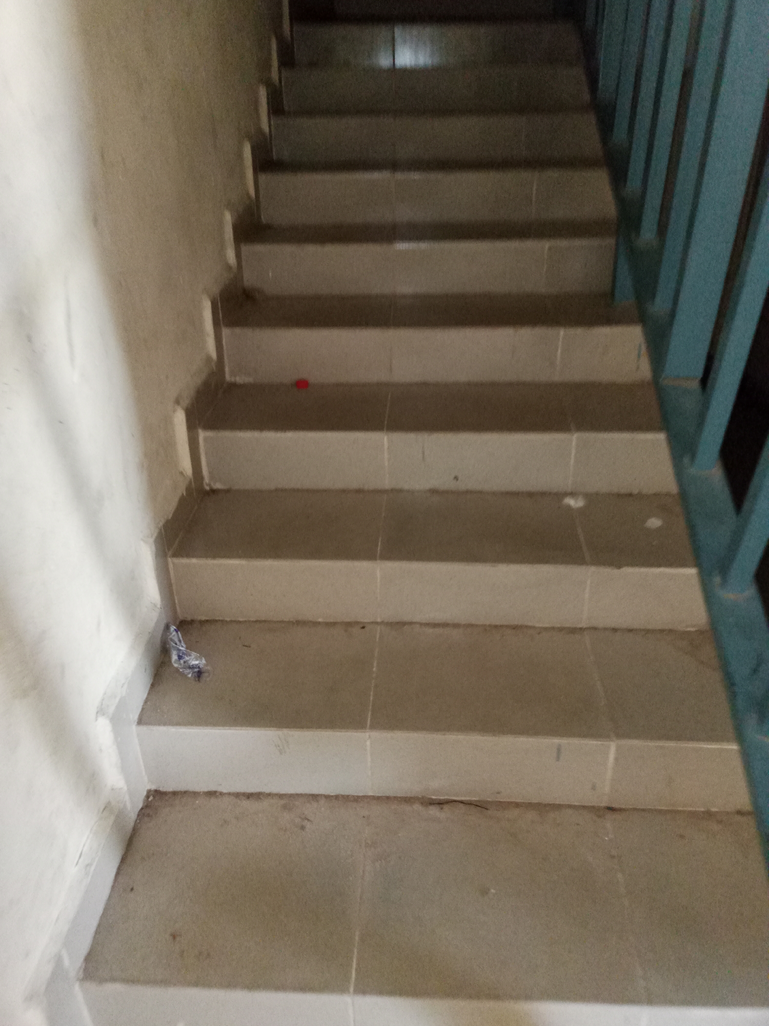 A close-up, low-angle shot shows a concrete staircase with light-colored tile risers and treads. A light blue metal railing runs along the right side of the stairs. On the wall to the left, there is a small piece of crumpled plastic debris. The scene is an interior staircase, likely within a building in Jalingo, Nigeria. The lighting is dim, suggesting an indoor environment without direct sunlight. The overall impression is of a functional, somewhat worn staircase.