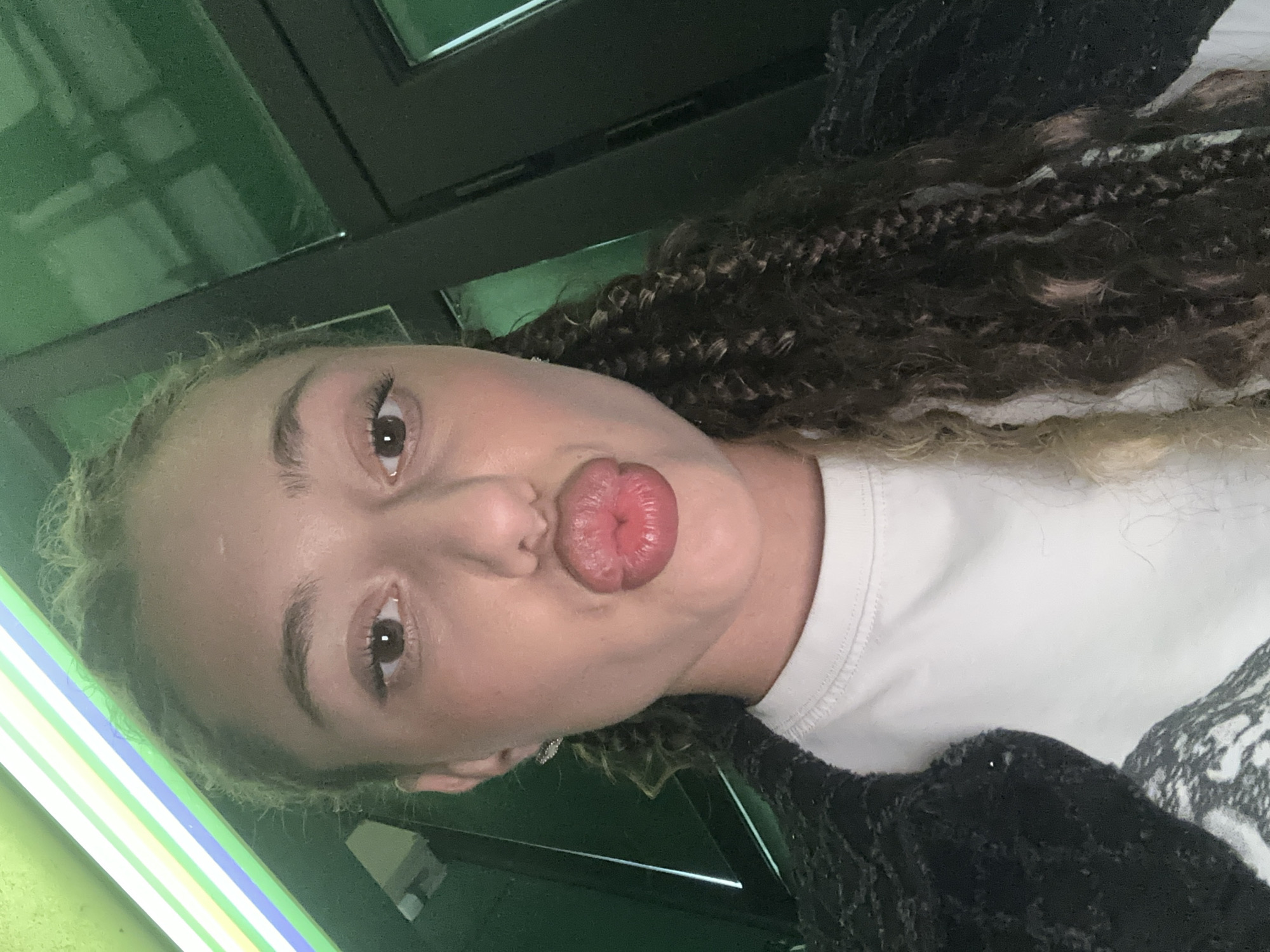 A close-up, selfie-style portrait of a young woman with curly, braided hair and prominent pouty lips, taken indoors with a green-tinted background. The woman is wearing a white t-shirt and a dark, textured jacket. The lighting suggests an artificial light source, possibly neon, with colored stripes visible at the top left of the frame. There are no discernible elements indicating a specific location within the city of Gabrovo, Bulgaria.