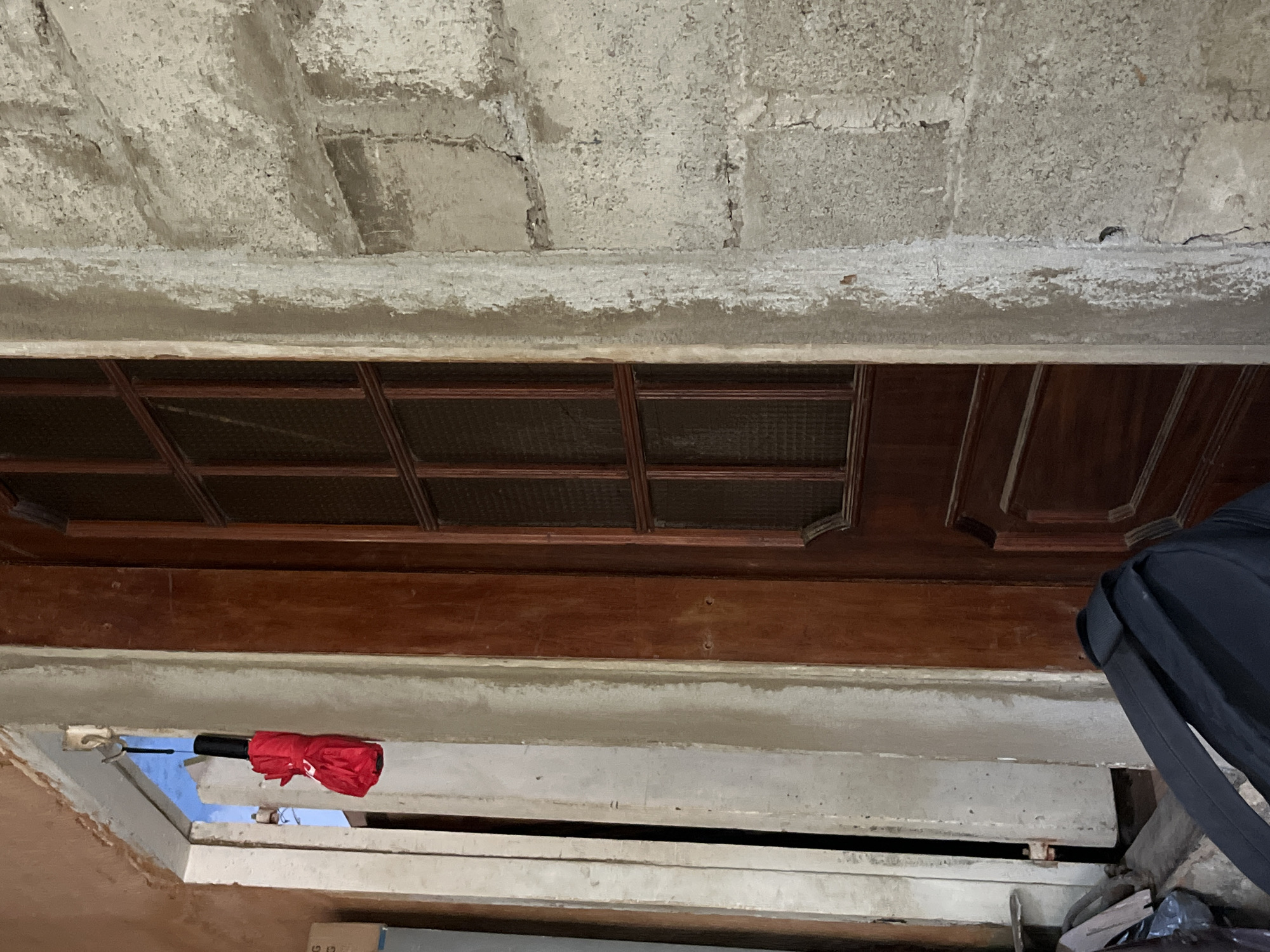 In Juan Griego, Venezuela, a red, folded umbrella hangs from a hook on a light-colored doorframe. To the right of the doorframe is a dark wooden door with a glass pane. To the left of the doorframe, a second doorframe is visible, with a black backpack resting on the floor in front of it. The wall to the right of the wooden door is made of concrete blocks. The overall scene appears to be an interior or semi-exterior space, possibly a doorway or entryway.