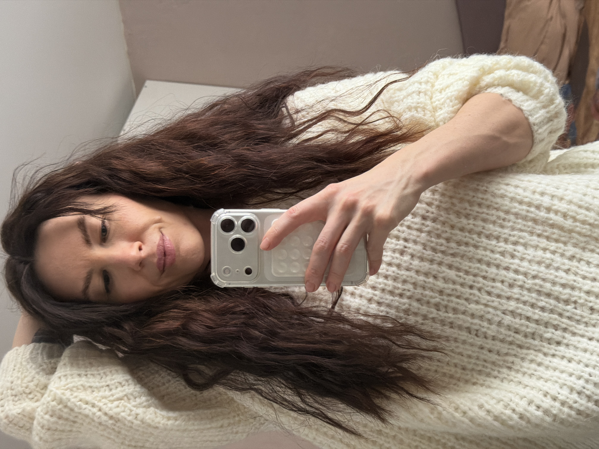 A female, approximately 25-35 years of age, is depicted from the upper torso upwards, taking a self-portrait indoors in Perm, Russia. She has long, dark brown, wavy hair and is wearing a cream-colored, thick-knit sweater. Her right hand holds a light-colored smartphone, equipped with a clear, textured protective case and a visible triple-lens camera array, positioned to capture her reflection. Her left arm is raised, with the hand partially obscured behind her head. Her gaze is directed downward, and her lips form a subtle, closed-mouth smile. The background features a plain white ceiling and an upper wall area, transitioning from light-colored on the left to a light beige or brown hue on the right, with a partial white vertical structure and blurred darker elements suggesting interior furnishings.