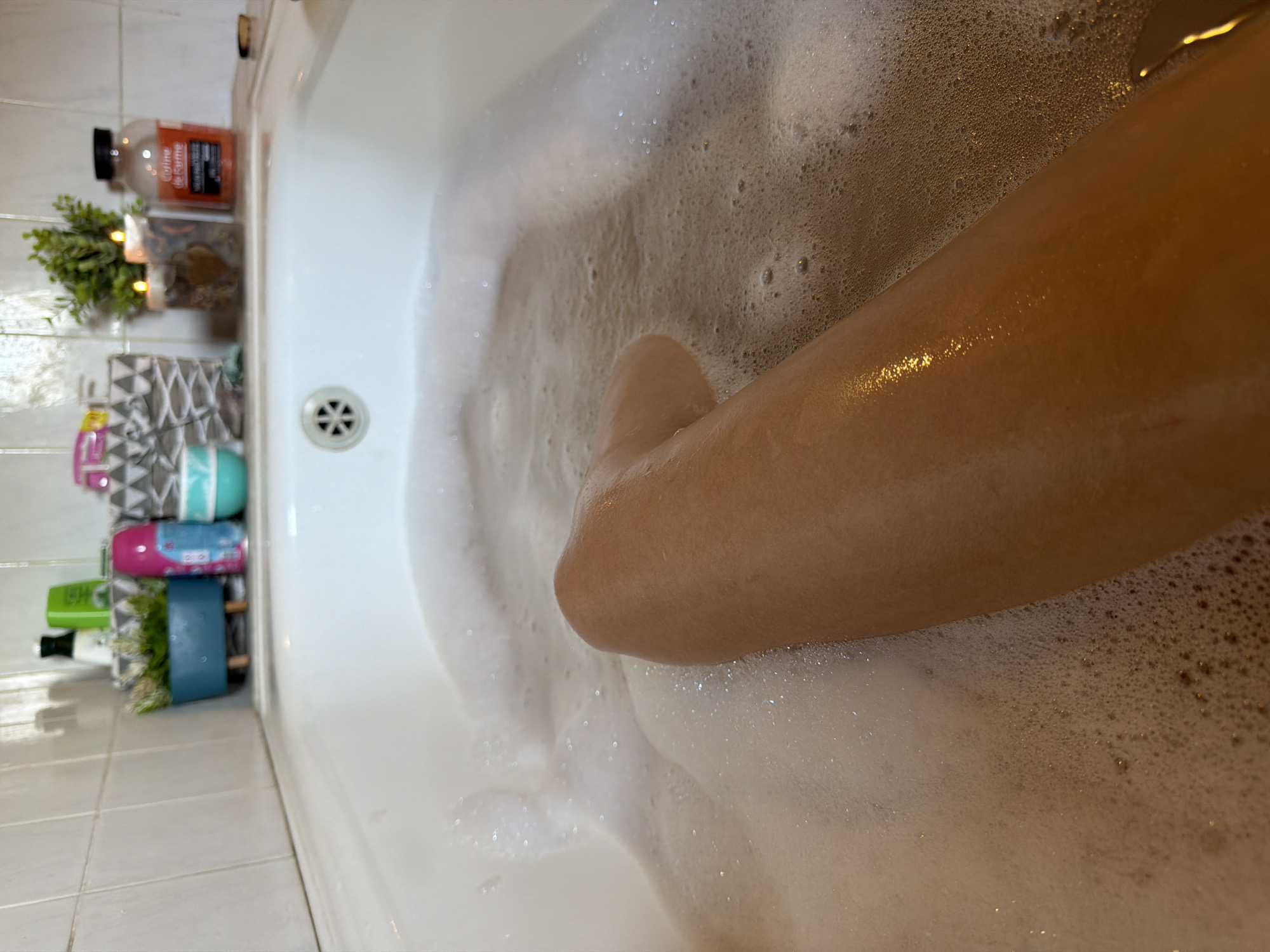 A close-up shot shows a person's leg submerged in a bathtub filled with foamy water. The leg, with smooth, tanned skin, is positioned diagonally across the frame, with the knee visible. Bubbles and foam cover the majority of the image, creating a soft, textured foreground. In the background, out of focus, are items on the edge of the bathtub, including bottles, a small plant in a pot, and candles. The tiling of the bathroom wall is also visible. The scene appears to be set in a bathroom, likely within a residential building.
