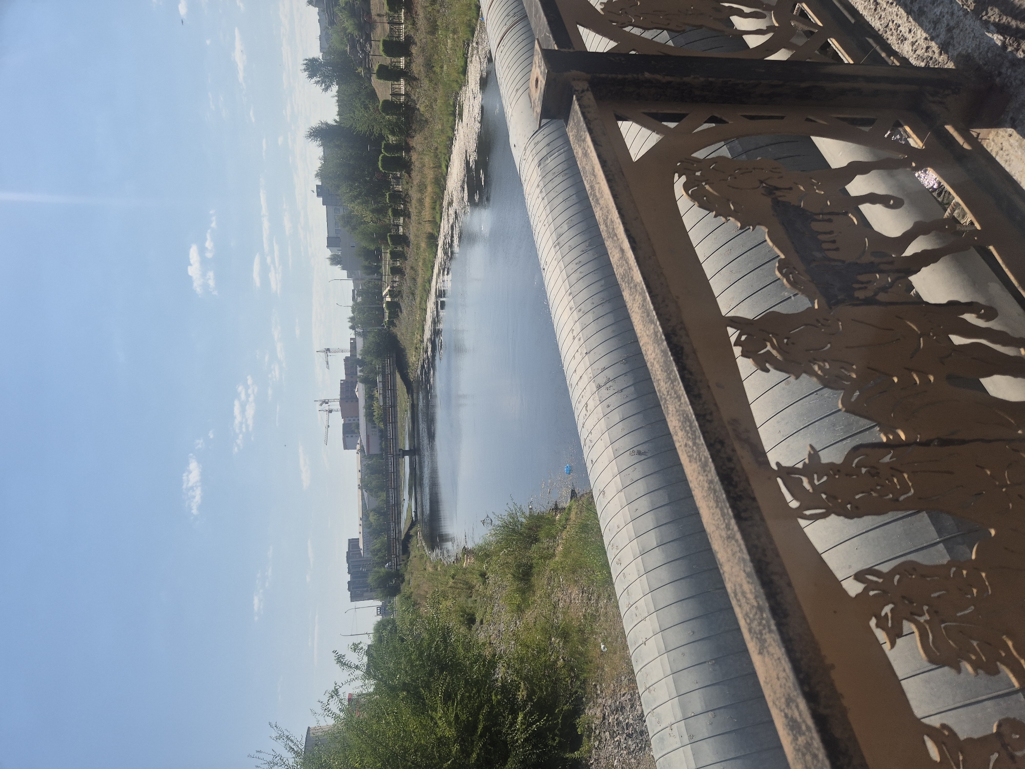The image shows a body of water, likely a river or canal, flowing through an urban environment. In the foreground, a decorative metal railing with cutouts of horses is visible, above a large, ribbed pipe. The railing appears weathered and rusty. The water is calm and reflects the blue sky, with some scattered debris and vegetation along its banks.
On the far bank, there are trees and bushes, followed by a street with buildings in the distance. Several multi-story buildings are visible, some appearing to be residential or commercial structures. Construction cranes are present in the background, suggesting ongoing development. A bridge crosses the water further in the distance.
The overall scene suggests a developed urban area with signs of both existing infrastructure and new construction. The presence of the decorative railing and the large pipe indicates a functional, possibly industrial or infrastructural aspect to the area, alongside the natural element of the water.
The image can be geolocated to Kokshetau, Kazakhstan, based on the specific architectural styles of the buildings and the general landscape, which are characteristic of the region. The style of the railing is also consistent with public art installations found in some Central Asian cities.