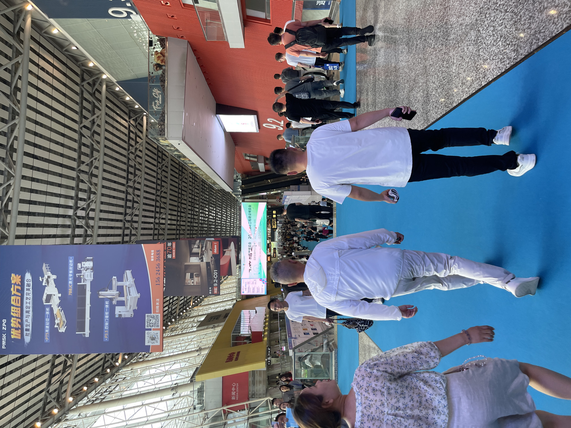 The image depicts an indoor exhibition hall, likely a convention center, in Haizhu District, China. The scene is filled with numerous people walking on a bright blue carpeted pathway. Overhead, large banners advertising various products and companies are suspended from the high, industrial-style ceiling structure. The banners feature images of machinery and text in Chinese. Along the right side of the walkway, a vibrant red wall rises, punctuated by circular design elements and large signage with the number "9.2". The overall atmosphere suggests a busy trade show or expo, with attendees moving through the exhibit space.