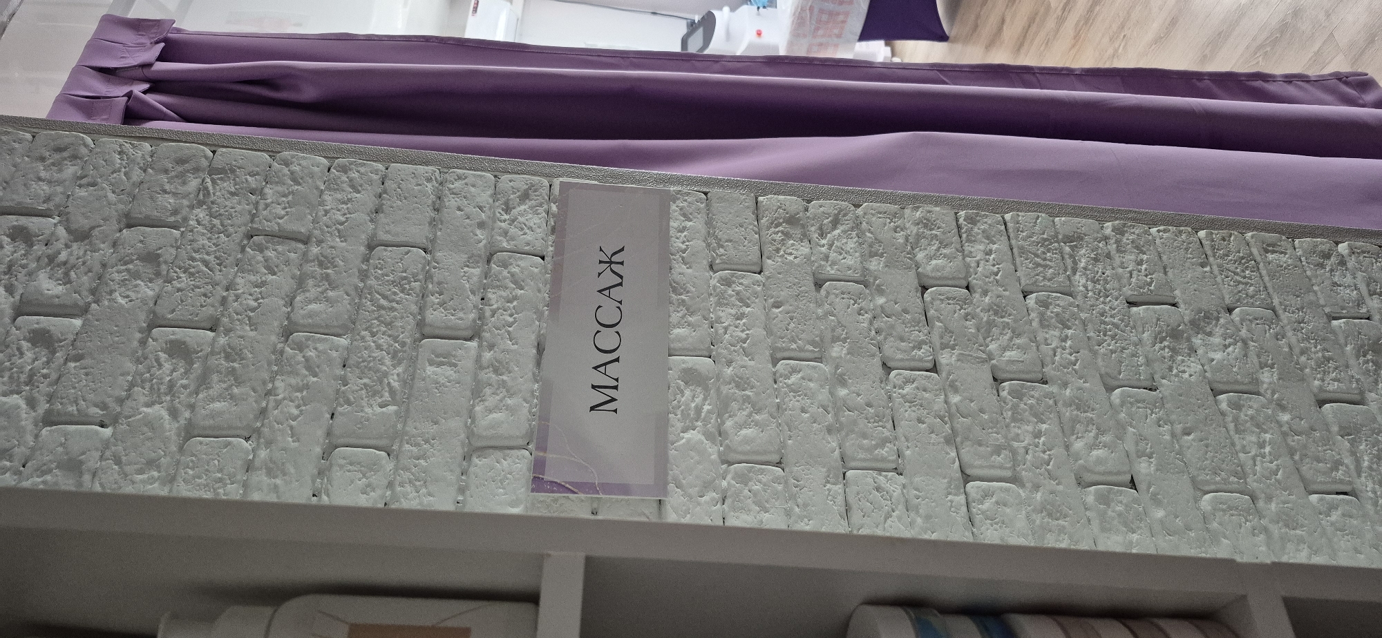 Here's a detailed description of the image:
The image features a close-up view of a white, textured brick-like wall. The bricks are arranged in an offset pattern and have a rough, uneven surface. A vertical sign with the word "МАССАЖ" (MASSAGE) in Cyrillic script is attached to the wall, partially obscuring the brick pattern.
Above the wall, a purple or lavender-colored fabric drapes down, giving the impression of a curtain or some form of textile covering.
At the bottom of the frame, a shelf-like structure is visible, with some objects on it. These include what appears to be a white or light-colored object partially visible in the bottom left corner and several cylindrical items in the bottom right.
The overall environment seems to be an interior space, possibly a spa, salon, or retail establishment. The location is unknown.