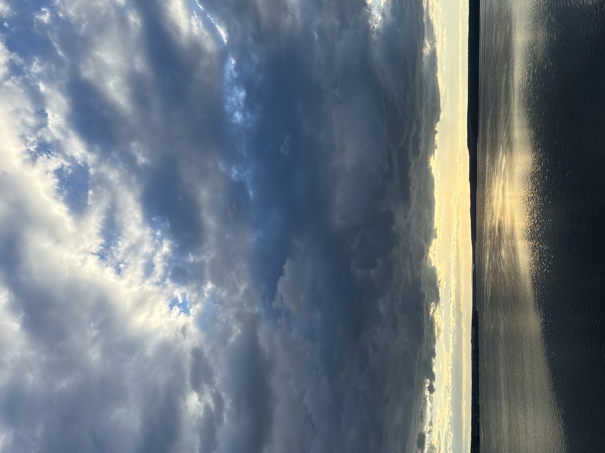 The image depicts a wide body of water, possibly a river or lake, under a dramatic sky filled with dark, heavy clouds. The water is calm, with gentle ripples reflecting the muted sunlight breaking through the clouds. A dark, forested shoreline is visible in the distance, with a hint of settlement along the coast. The overall mood is one of impending weather change, with the dark clouds contrasting with the slivers of bright sky and reflected light on the water. The specific location in Votkinsk, Russia, is not visually identifiable from the image alone, but the scene suggests a natural landscape with a distant human presence. There are no people or specific objects clearly visible in the frame.