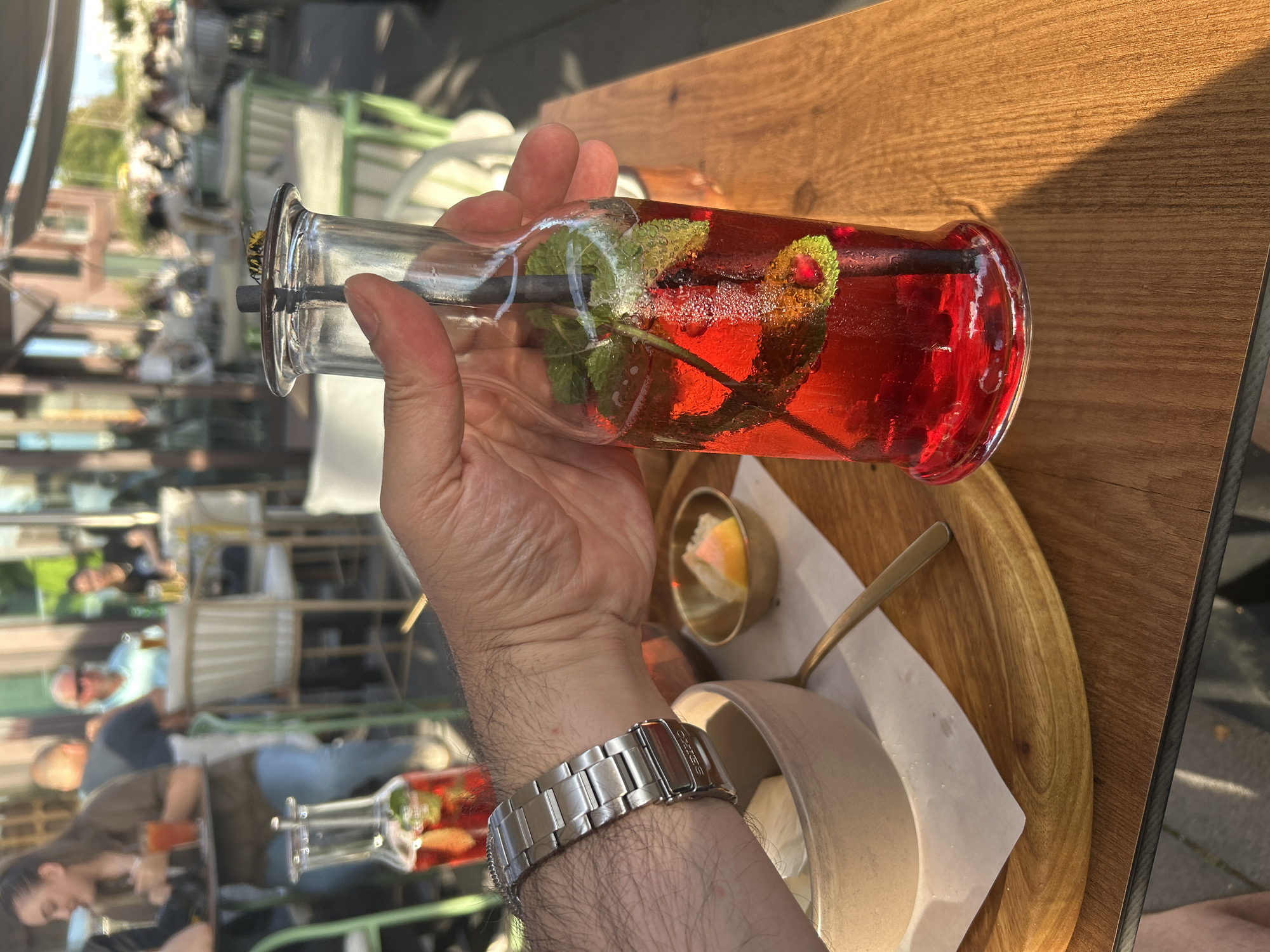 A close-up shot focuses on a glass bottle filled with a red, cold beverage and garnished with mint and possibly berries, held by a person's hand wearing a silver watch. The bottle is resting on a wooden table at an outdoor cafe in Berlin, Germany. In the background, several people are seated at tables, some dining and others conversing, beneath umbrellas, suggesting a pleasant daytime setting. The ambiance is relaxed, typical of a European city during warm weather.