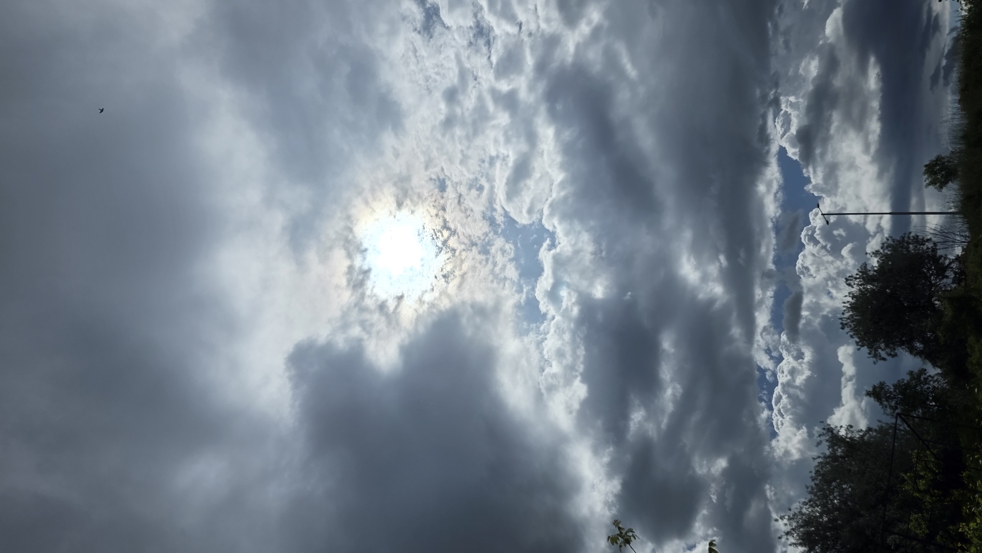 The image shows a cloudy sky with the sun shining through a break in the clouds. The lower right portion of the image shows a tree line, with a utility pole visible. The image appears to have been taken outdoors, although no specific information about the location or whether it is in Blagoevgrad, Bulgaria, can be determined from the given information. There are no people or actions visible.