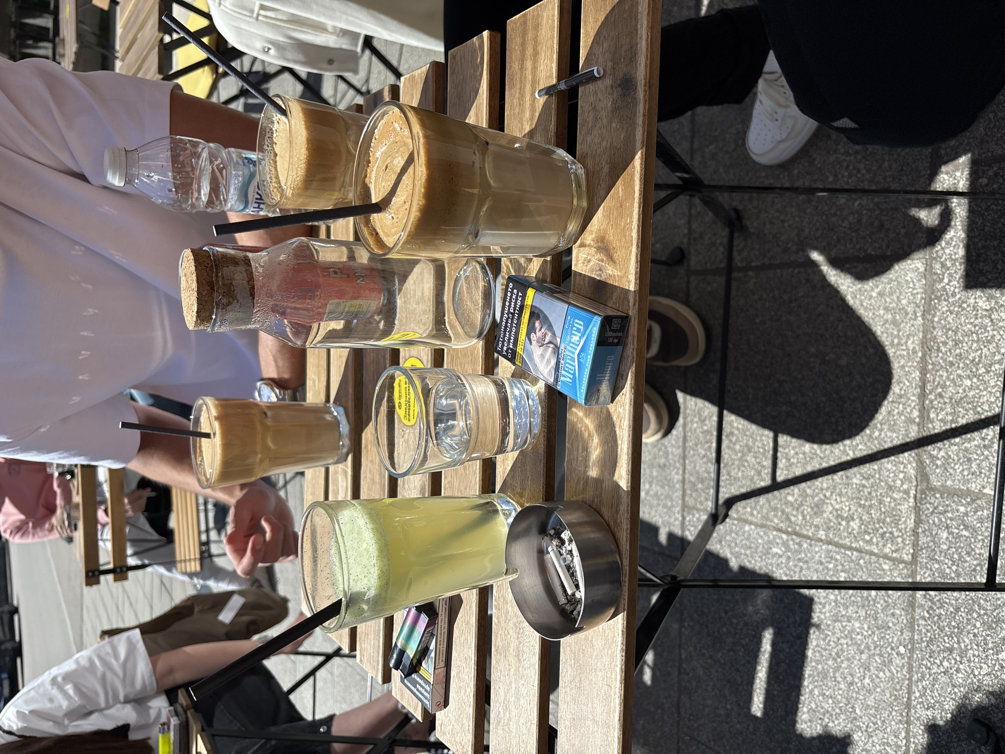 The image captures an outdoor scene, likely at a café or similar establishment. The main focal point is a wooden table displaying an assortment of drinks, including what appear to be coffee beverages, a lemonade-type drink, a bottle of water, and an unidentified bottle with a cork. Also on the table are a pack of Marlboro cigarettes, an ashtray with cigarette butts, and a few other items. The image provides a top-down view of the table's arrangement and the legs of several individuals seated around it. The location is not explicitly mentioned in the image, and information about Gabrovo, Bulgaria, cannot be confirmed solely from the visual context provided.