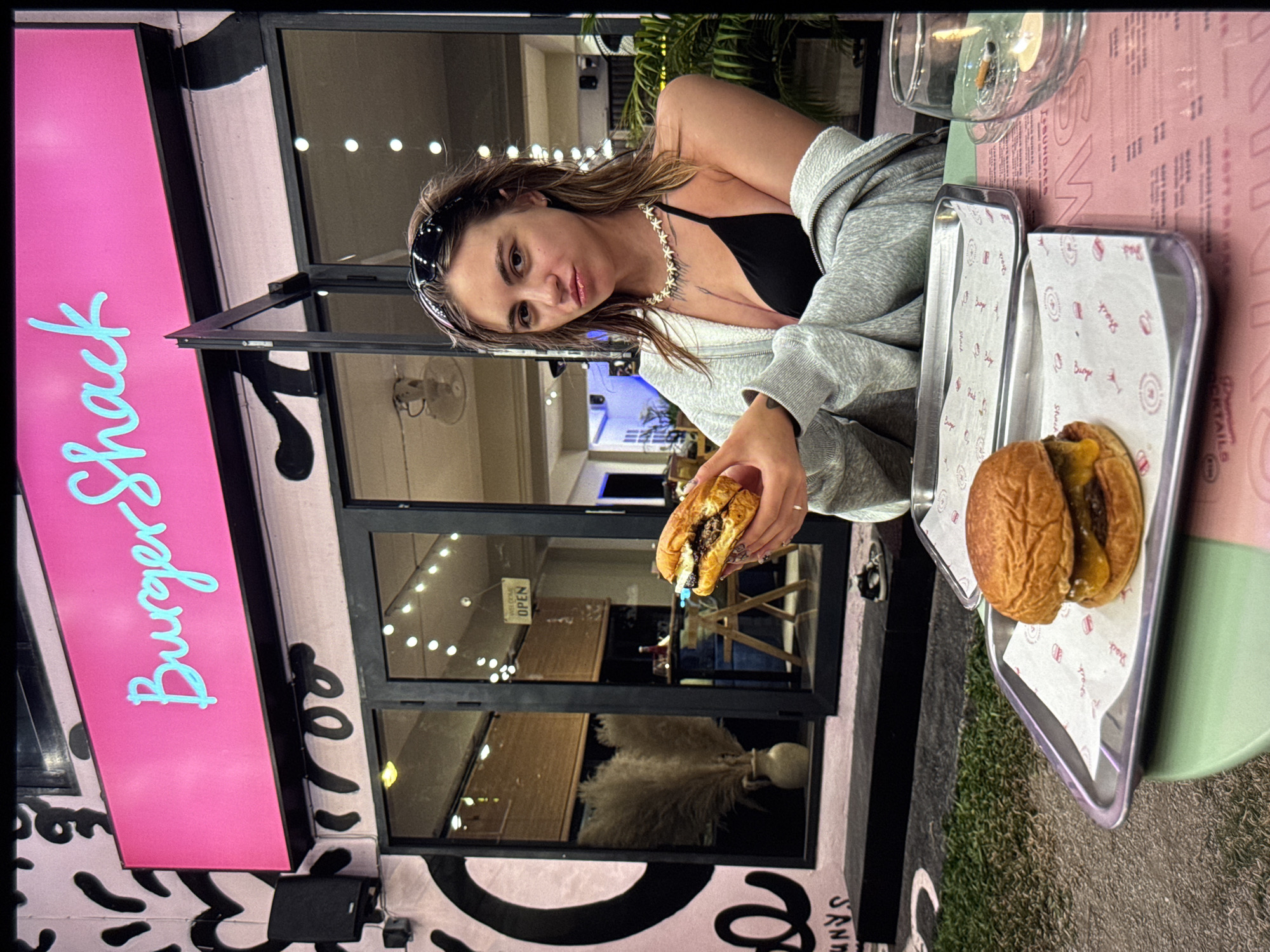 A young woman with wet, shoulder-length hair and a bandana on her head is sitting at a table outside a restaurant. She is wearing a black bikini top and a grey hooded cardigan. She is holding a burger in her right hand and looking at the camera with a pouty expression. In front of her on the table are two metal trays, each holding a burger. The restaurant behind her has a bright pink sign that says "Burger Shack" in blue script. The building has large glass windows with string lights visible inside. The overall scene suggests an outdoor dining experience at a casual burger joint.