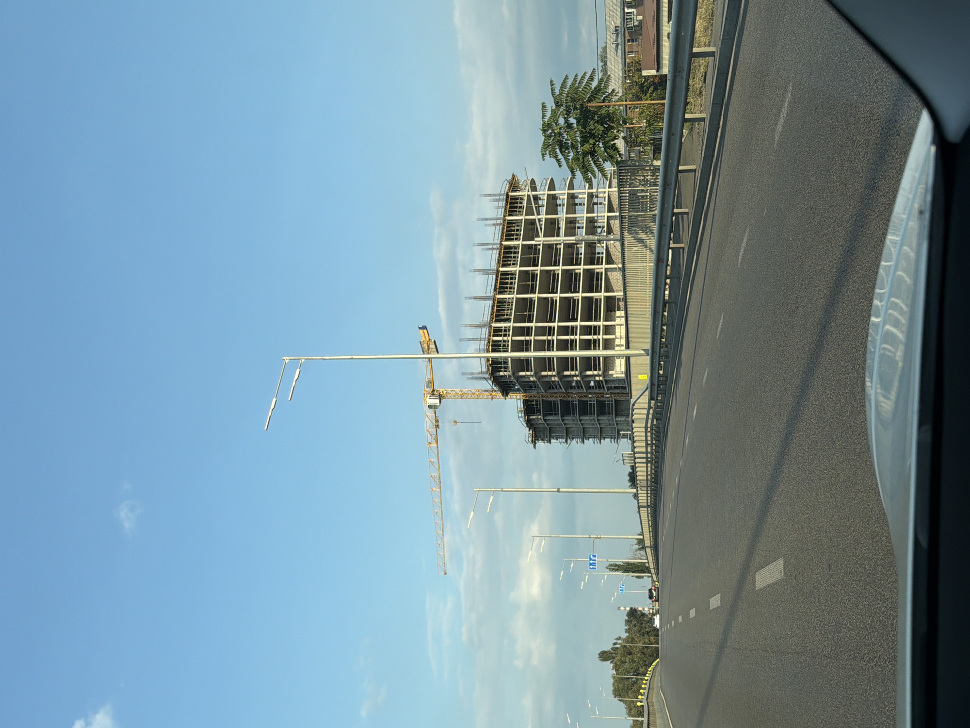 The image depicts a view from a vehicle driving on a road, likely a bridge, in Kyiv, Ukraine. In the background, a large, unfinished multi-story building is under construction. A yellow tower crane is positioned over the building, suggesting ongoing construction activity. The sky is a clear blue with some scattered clouds. Streetlights line the sides of the road. Several cars are visible on the road ahead, indicating traffic. The road has white lane markings and a metal guardrail separates the road from an embankment on the right. Trees and other buildings can be seen in the distance to the left and right of the construction site. The perspective suggests the photo was taken during the daytime.
