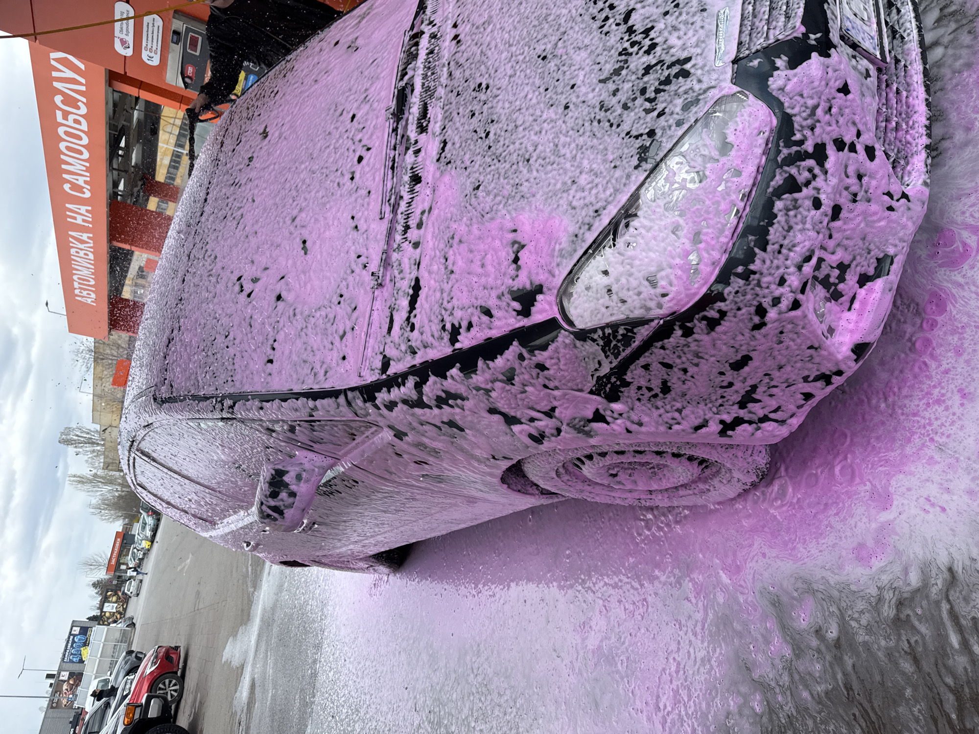 The image depicts a black car undergoing a wash at what appears to be a self-service car wash facility. The car is heavily covered in pink soap foam. The car's front headlight, hood, windshield, and front tire are clearly visible beneath the foam. Water and soapy residue are running off the car and onto the pavement. In the background, there's car wash signage with text in Cyrillic, suggesting the location is in a country that uses this alphabet. Vehicles are parked in the lot behind the carwash station. Based on the sign "АВТОМИВКА НА САМООБСЛУЖВАНЕ," this is likely a car wash facility, and based on the text on the sign, it seems to be located in Sofia, Bulgaria. A person is holding the washing apparatus.