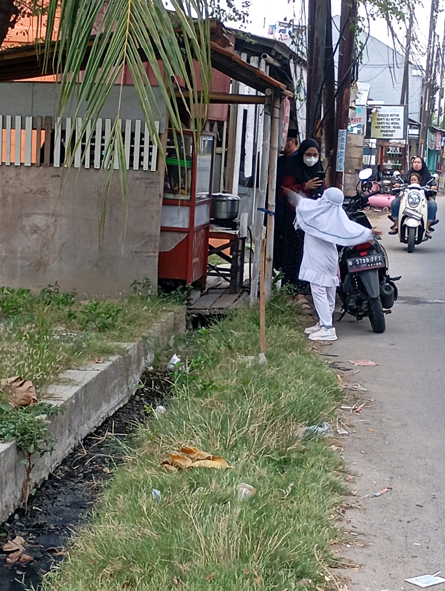 In Babelan, Indonesia, a scene unfolds on a roadside. A woman in a black hijab and a white mask stands with her phone, reaching out. Beside her, a child in a white hijab and matching pants stands with arms outstretched, facing a black motorcycle. Another motorcycle, white and with two riders, is parked on the road to the right. In the background, a small food stall with a red frame is visible, and beyond that, buildings and power lines. The foreground features a concrete drainage ditch filled with dark water and surrounded by overgrown green grass, with scattered debris on the side of the road.