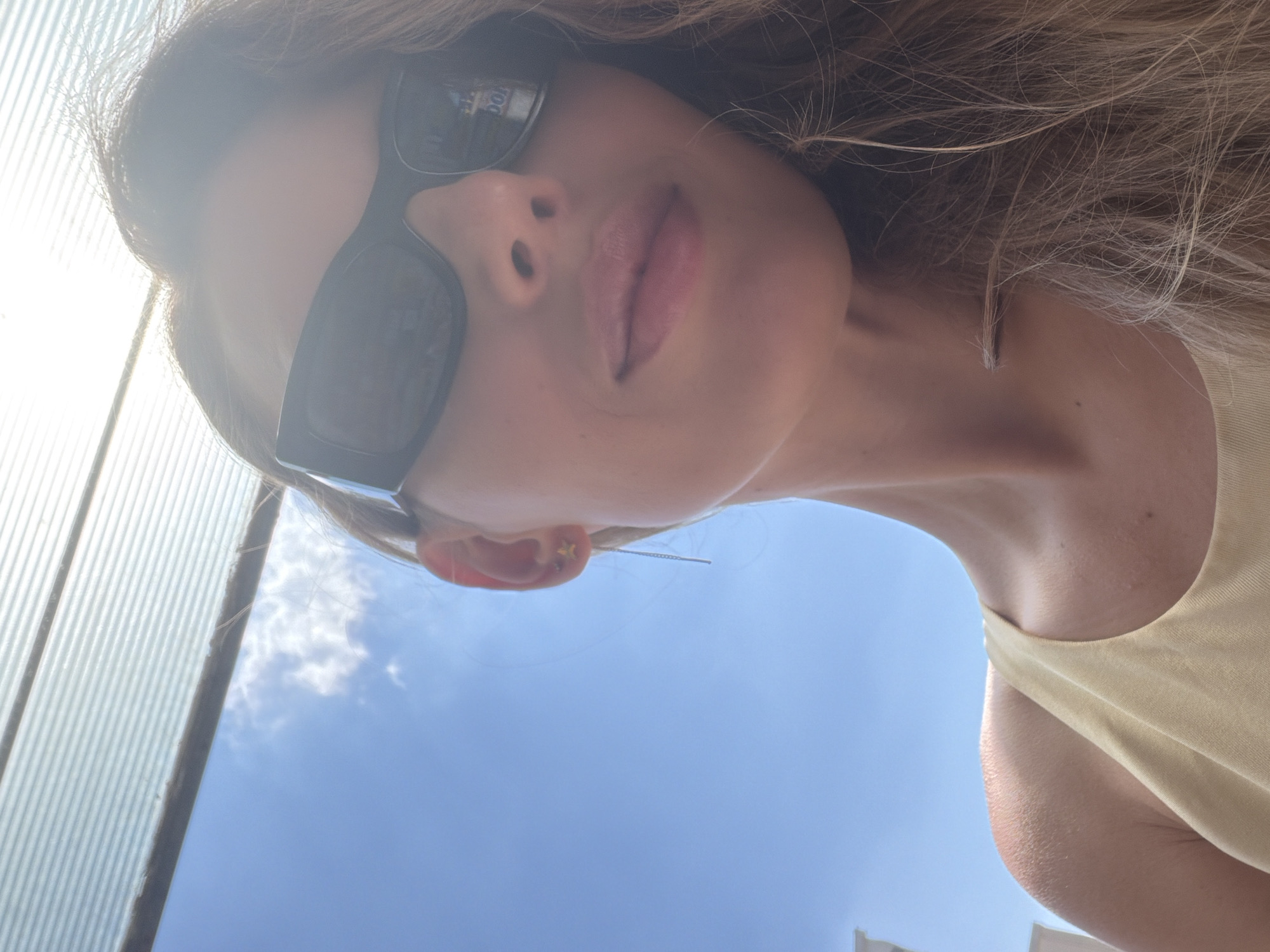 A woman wearing sunglasses and a light-colored tank top is captured in a selfie with a bright blue sky and some clouds in the background. She has long, wavy blonde hair and a small star-shaped earring in her visible ear. The image appears to be taken outdoors, possibly on a sunny day. The surrounding environment is not clearly identifiable as Budva, Montenegro, but the scene suggests a warm, possibly coastal, location given the bright sky.