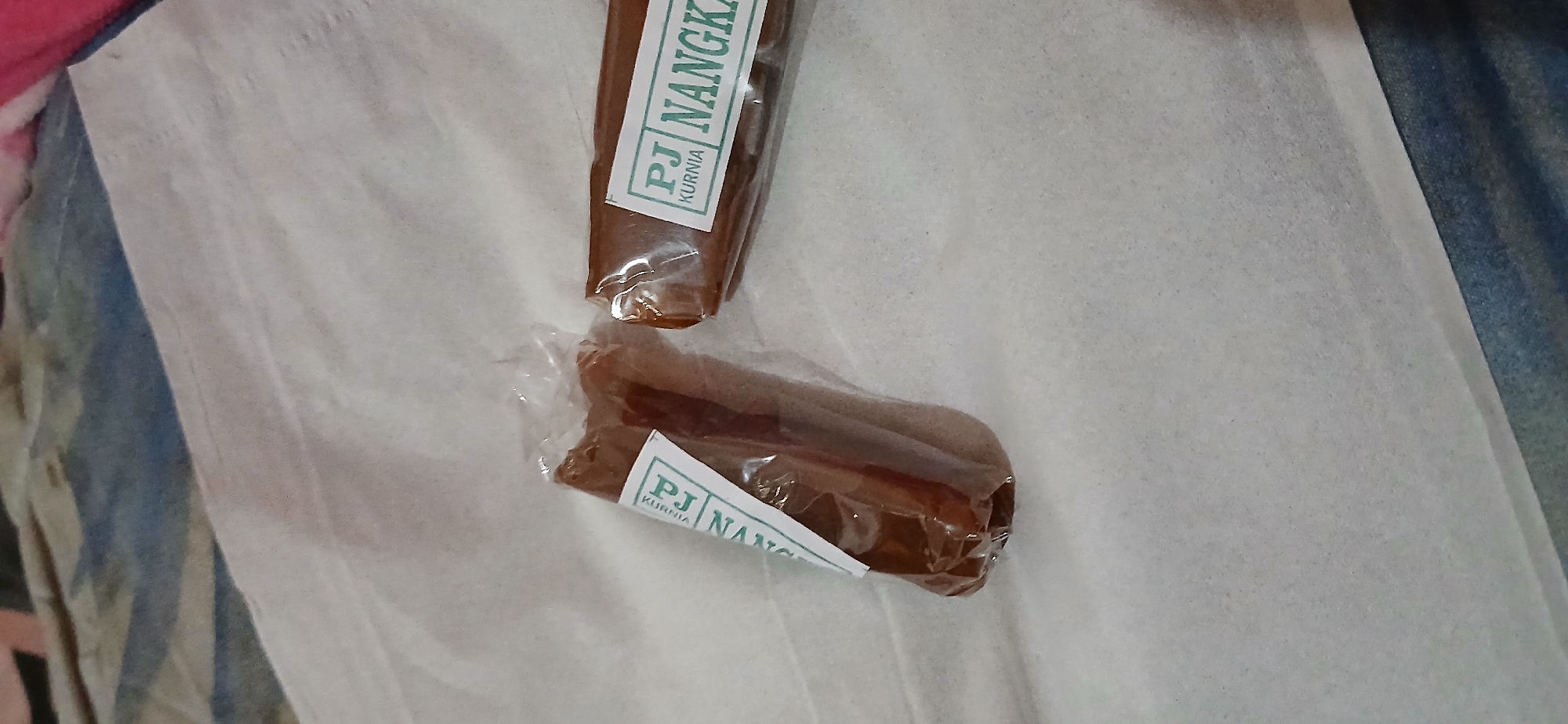 The image shows two individually wrapped pieces of what appears to be candy or confectionary, lying on a white textured surface. Each piece is elongated and cylindrical, wrapped in clear plastic. A white label with green text is affixed to each wrapper. The text on one label clearly reads "PJ NANGKA" and "KURNIA." The other label is partially visible and appears to have similar text. The background is a blurred mix of fabric textures, with a hint of blue and patterned fabric. The lighting is diffuse, and the image is taken from a slightly elevated angle, looking down. There are no people or explicit environmental cues suggesting a specific location within Samarinda, Indonesia.