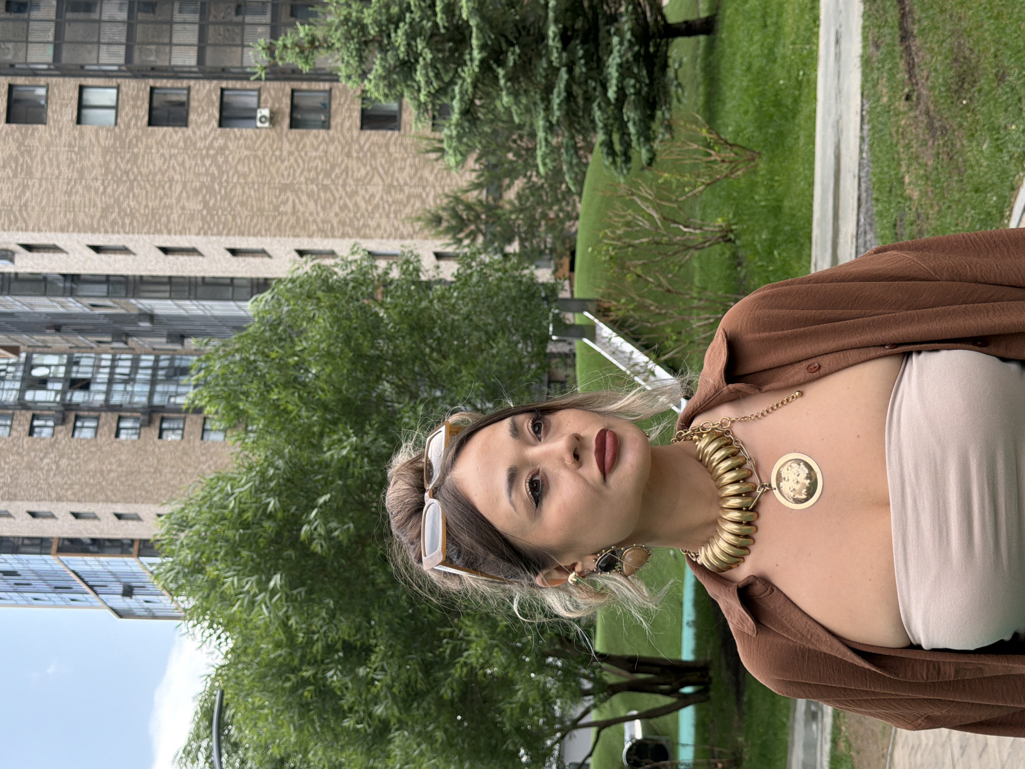 A woman with her hair pulled up and sunglasses perched on top of her head stands outdoors in front of a multi-story building. She is wearing a brown, open-collared shirt over a strapless, beige top and a large, gold statement necklace with a round pendant. She has dramatic eye makeup and dark lipstick. The background features lush green trees, manicured grass, and a modern brick building with many windows, characteristic of urban residential areas such as those found in the city of Krasnoyarsk, Russia. A metal slide structure is visible on the left side of the frame, partially obscured by trees.