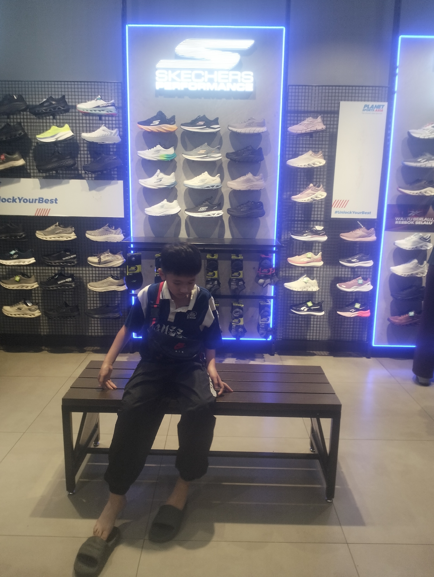 A young male is sitting on a wooden bench in a Skechers store in Surabaya, Indonesia. He is wearing a blue and white striped collared shirt with dark pants and dark sandals. Behind him, on display shelves, are numerous pairs of Skechers shoes. The store features bright blue neon lighting behind the shoe displays and the Skechers logo. The floor is tiled. The overall environment suggests a retail space dedicated to athletic footwear.
