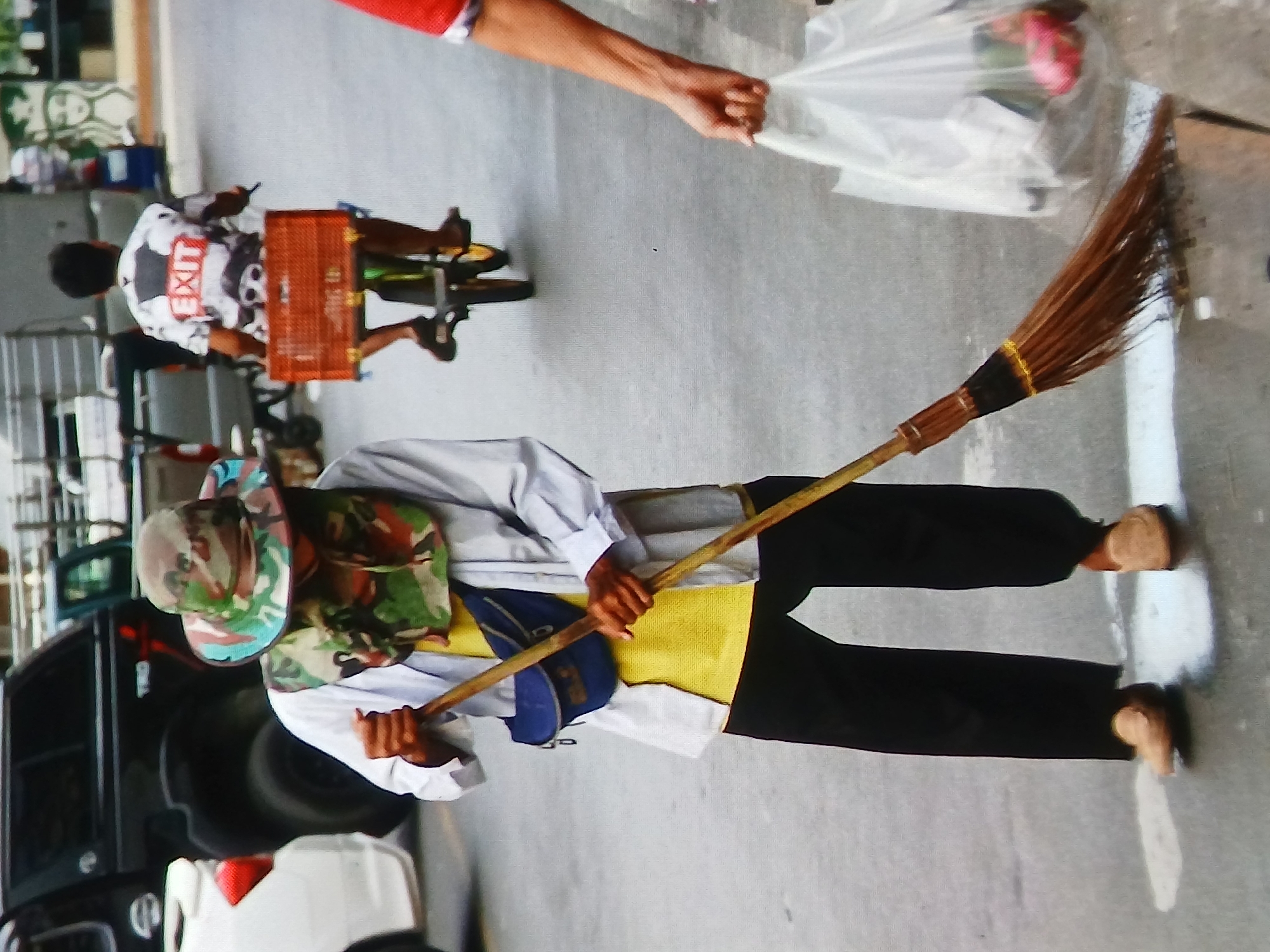 In a city street in Setia Bakti, Indonesia, a person wearing a camouflage hat, a white long-sleeved shirt over a yellow t-shirt, black pants, and tan loafers sweeps the ground with a broom. A blue fanny pack is worn across their chest. In the background, another person rides a bicycle with a red basket attached to the back, wearing a white shirt with "EXIT" emblazoned on it in red. A white plastic bag, containing red fruit, is held by a third person partially visible on the right side of the frame. Several vehicles, including a black pickup truck and a white car, are parked on the street. The overall scene depicts an urban street environment with everyday activities taking place.