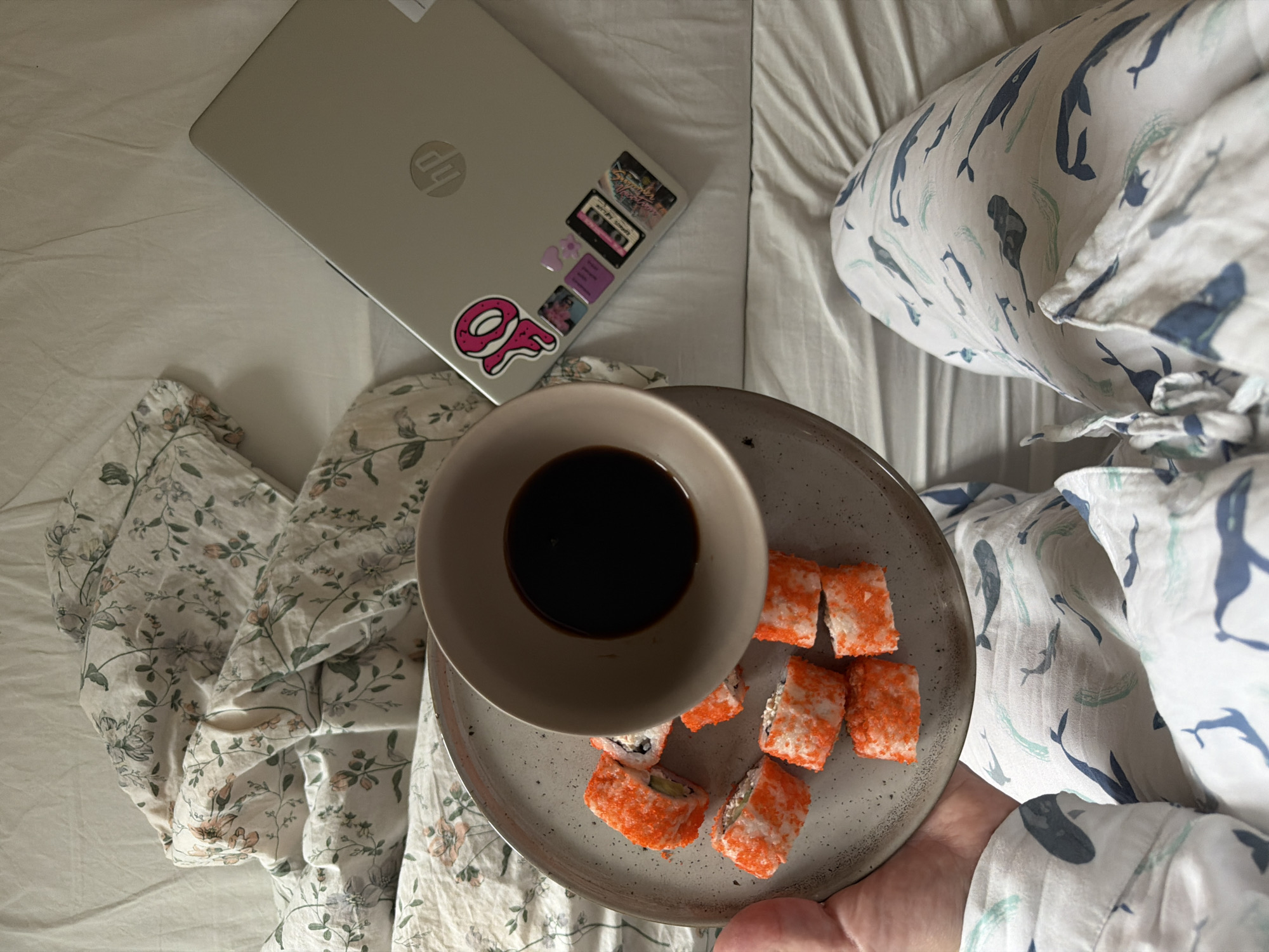 The image shows a top-down view of a person in bed with a laptop and a plate of sushi. The laptop, a silver HP model adorned with stickers, sits on the left side of the frame. A round gray plate holds several pieces of sushi covered in orange roe and a bowl of dark soy sauce. The person is wearing pajamas with two different patterns: one side features a floral print, and the other has a whale design. The scene is set on a white bed with wrinkled sheets. The specific location in Izhevsk, Russia, cannot be determined from the image alone.