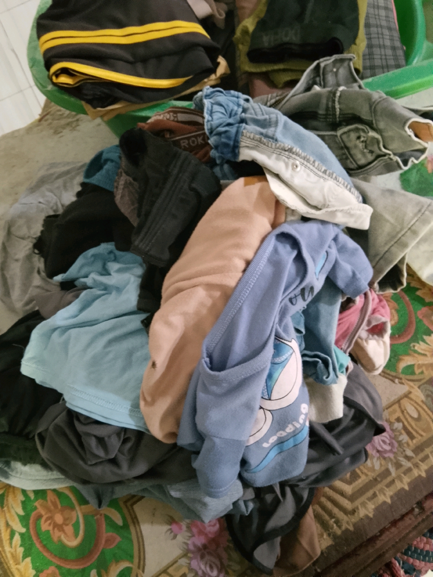 The image shows a pile of laundry in various colors and fabrics, including a black and yellow striped bag, denim jeans, a pink top, and several t-shirts in shades of blue, gray, and peach. The laundry is situated on a patterned rug, with some items spilling out of a green plastic container. The scene appears to be indoors, with a tiled floor visible in the background. The lighting is dim, suggesting it might be nighttime or an indoor setting with limited natural light. There are no people or animals visible in the image. The overall impression is one of a domestic scene, possibly a laundry room or bedroom, in Karang Gading, Indonesia.