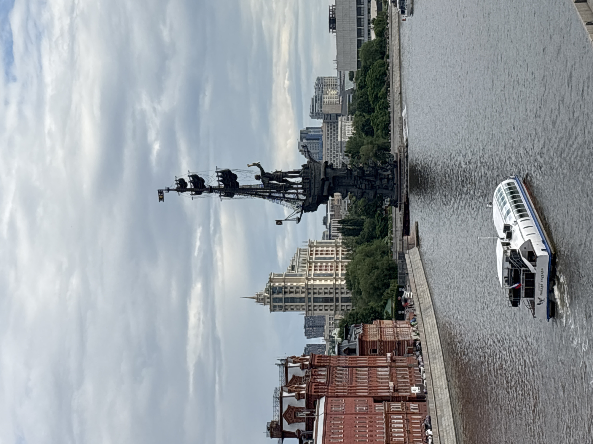 The image depicts a cityscape, presumably Moscow, Russia, given the presence of the Peter the Great Statue. The monument is the focal point, featuring figures atop a tall ship. The Moscow River runs vertically in the image, with a passenger boat visible on its surface. Various buildings and structures line the riverbanks, including red-brick historical buildings and modern high-rises. The sky is overcast with numerous clouds. The image seems to be taken from an elevated position, providing a comprehensive view of the river and its surroundings.
