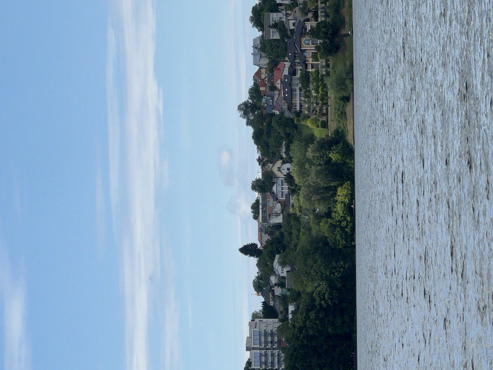 The image displays a panoramic view of a waterfront residential area situated on a hillside overlooking a body of water. The scene is dominated by the blue sky with scattered clouds, the green foliage of trees and bushes, and a collection of houses of varying architectural styles and sizes nestled into the slope. In the foreground, the water's surface is textured with ripples, suggesting a gentle breeze.
Along the hillside, several prominent structures are visible. On the left side, a modern, multi-story building with a light-colored facade and large windows stands out. To its right, a cluster of smaller residential buildings, some with red-tiled roofs, are interspersed with trees. Further to the right, larger, more elaborate houses with well-maintained landscaping are present, some featuring decorative elements and multiple levels. A small white structure with a cross on top, possibly a church or chapel, is visible among the trees and buildings. The density of the greenery suggests a developed area with mature trees and carefully tended gardens.
The overall impression is one of a serene, established residential neighborhood located in a natural setting with access to water. The image is likely taken from a boat or the opposite bank of the water body, providing a distant perspective of the hillside community. The scene is devoid of any visible human activity in the immediate foreground or midground, lending a tranquil atmosphere. The location is described as being in the city of Ternopil, Ukraine, suggesting this is a view of one of its lakeside neighborhoods.