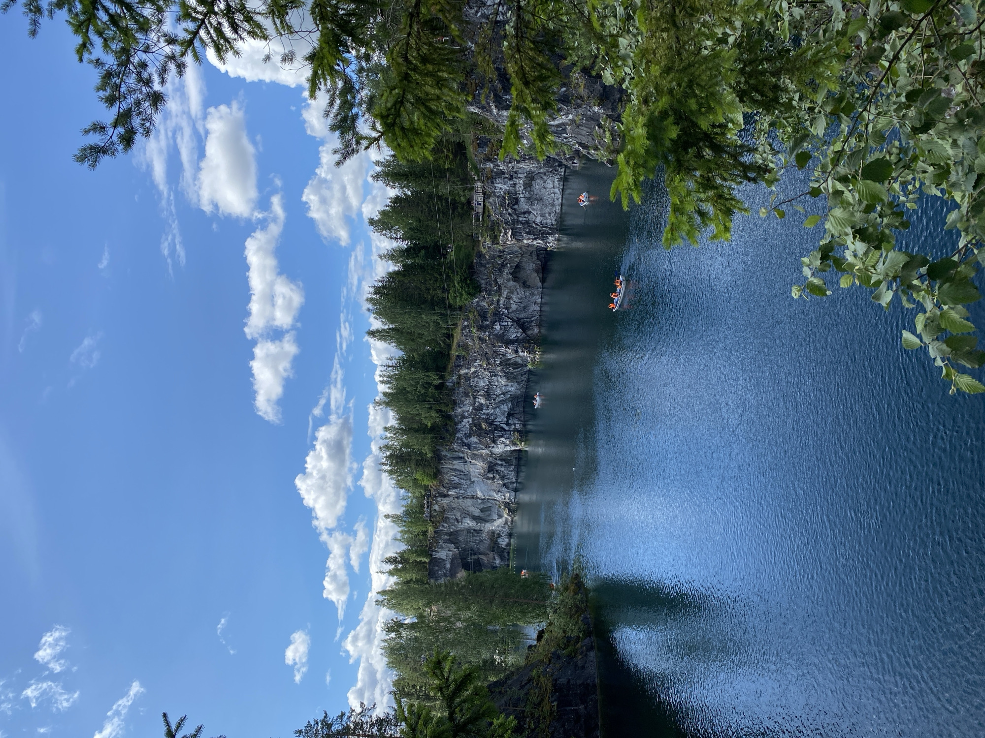 The image depicts the Marble Canyon in Ruskeala, Russia, a popular tourist destination known for its stunning natural beauty. The canyon features a deep, turquoise-blue lake surrounded by towering marble cliffs, indicative of its geological origins as a former marble quarry. Lush green trees and coniferous forests line the tops of the cliffs, creating a vibrant contrast with the cool tones of the water and stone.
Several small boats, likely rented by tourists, are visible on the lake, some carrying a few people wearing orange life vests. These boats are scattered across the water, suggesting a leisurely recreational activity. The sky above is a bright, clear blue, dotted with fluffy white clouds, indicating fair weather. The overall scene conveys a sense of tranquility and natural wonder, attracting visitors to explore the unique landscape.