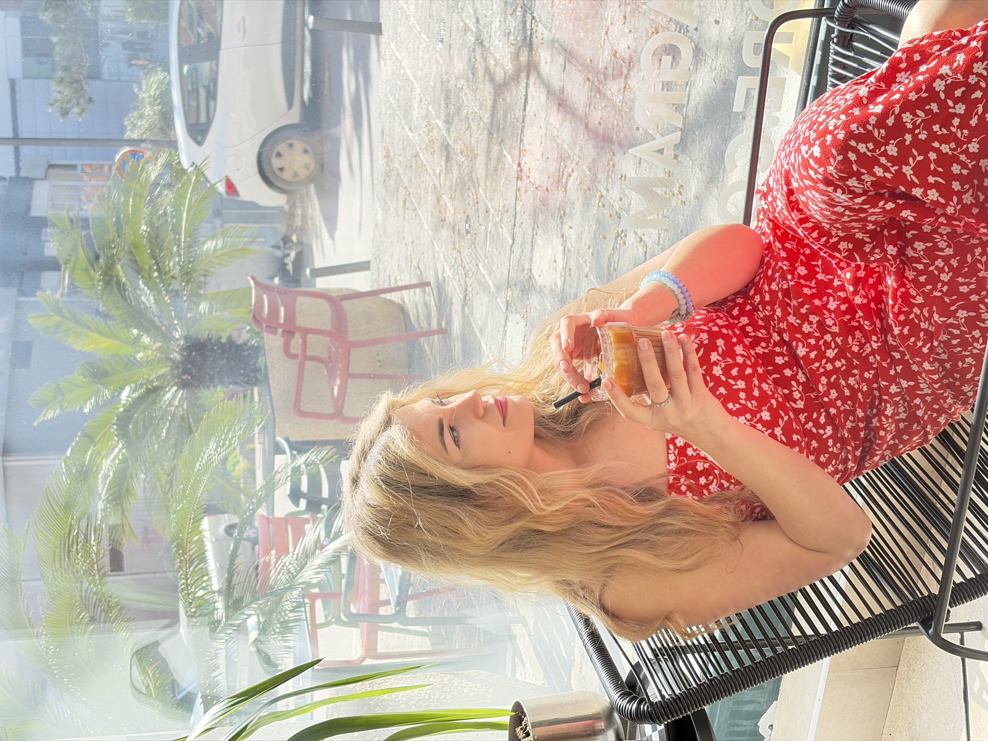 A blonde female individual is seated in a black slatted chair inside an establishment. She is wearing a red strapless dress adorned with a white floral pattern. Her left hand holds a clear glass containing an amber-colored beverage with a black straw. Multiple beaded bracelets, including blue, purple, and white beads, are visible on her left wrist, and a ring is on her left ring finger. Her gaze is directed towards the right. A green potted plant in a metallic container is situated to her left.
The scene is viewed through a large window, revealing an urban outdoor environment. Palm trees are prominently visible, reflecting sunlight. An outdoor seating area features several red metal chairs and a dark-colored chair. A white hatchback vehicle is visible on a paved street or parking area. The pavement consists of light-colored tiles or cobblestones. A red and white circular traffic sign is partially visible, along with two dark bollards. The inverted text "GRAM" is reflected on the lower right of the window, indicating the establishment is likely "Gram Cafe Budva," situated on Mediteranska Street in Budva, Montenegro.