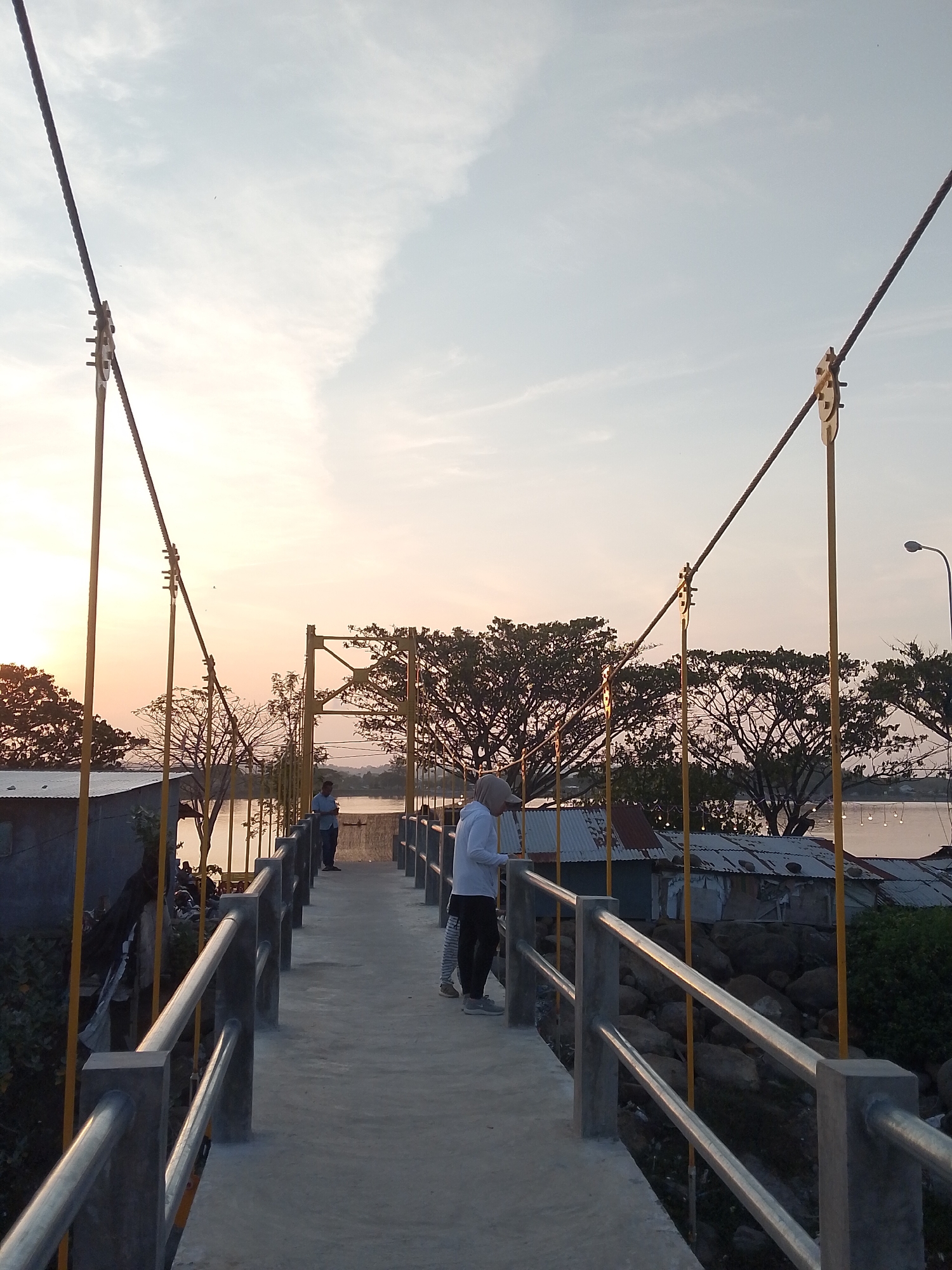 The image depicts a suspension bridge in Lhokseumawe, Indonesia, at what appears to be sunset. The bridge, constructed with grey concrete for the walkway and railings, is held by yellow metal supports and dark cables. Several people are visible on the bridge: one individual stands closer to the camera wearing a white long-sleeved shirt, dark pants, and a baseball cap, seemingly looking at something in their hands. A child stands next to them. Another person is visible further down the bridge.
On both sides of the bridge, there are structures that appear to be small buildings or houses with corrugated metal roofs. Large trees are silhouetted against the sunset sky. The presence of water suggests the bridge spans a river or lake. A streetlight is visible to the right. The sky exhibits a gradient of colors, ranging from a light blue to a warm orange at the horizon.