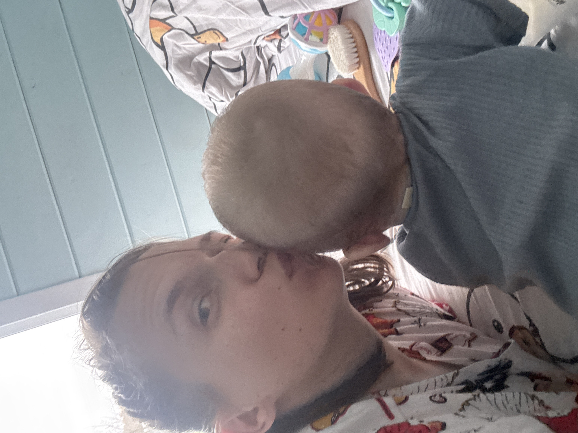 This image captures a tender, close-up moment between an adult and a baby in what appears to be an indoor setting in Razdolye, Russia. The perspective suggests a selfie or a photograph taken from above.
The adult, visible on the left and bottom of the frame, has light brown hair and a scattering of freckles on their face. Their eye is visible looking towards the camera, and their lips are slightly pursed, seemingly receiving or giving affection. They are wearing a light-colored top, possibly with a red pattern, matching a blanket or sheet that they are lying on.
The baby, seen from the top of their head and back, has very fine, light-colored hair and is wearing a gray, ribbed long-sleeved garment. Their head is gently pressed against the adult's face, near the mouth and nose, indicating a loving interaction like a kiss or a nuzzle.
The background reveals a light blue paneled wall on the left. On the right, behind the baby, several items associated with infants are visible on a white patterned blanket, which features cartoon animal figures such as a deer or reindeer. These items include a wooden-handled baby brush with soft bristles and a colorful rattle toy. The lighting is bright, suggesting natural daylight.
The image conveys a sense of warmth, intimacy, and a loving bond between the two individuals in a domestic environment. There is no visible text in the media file.