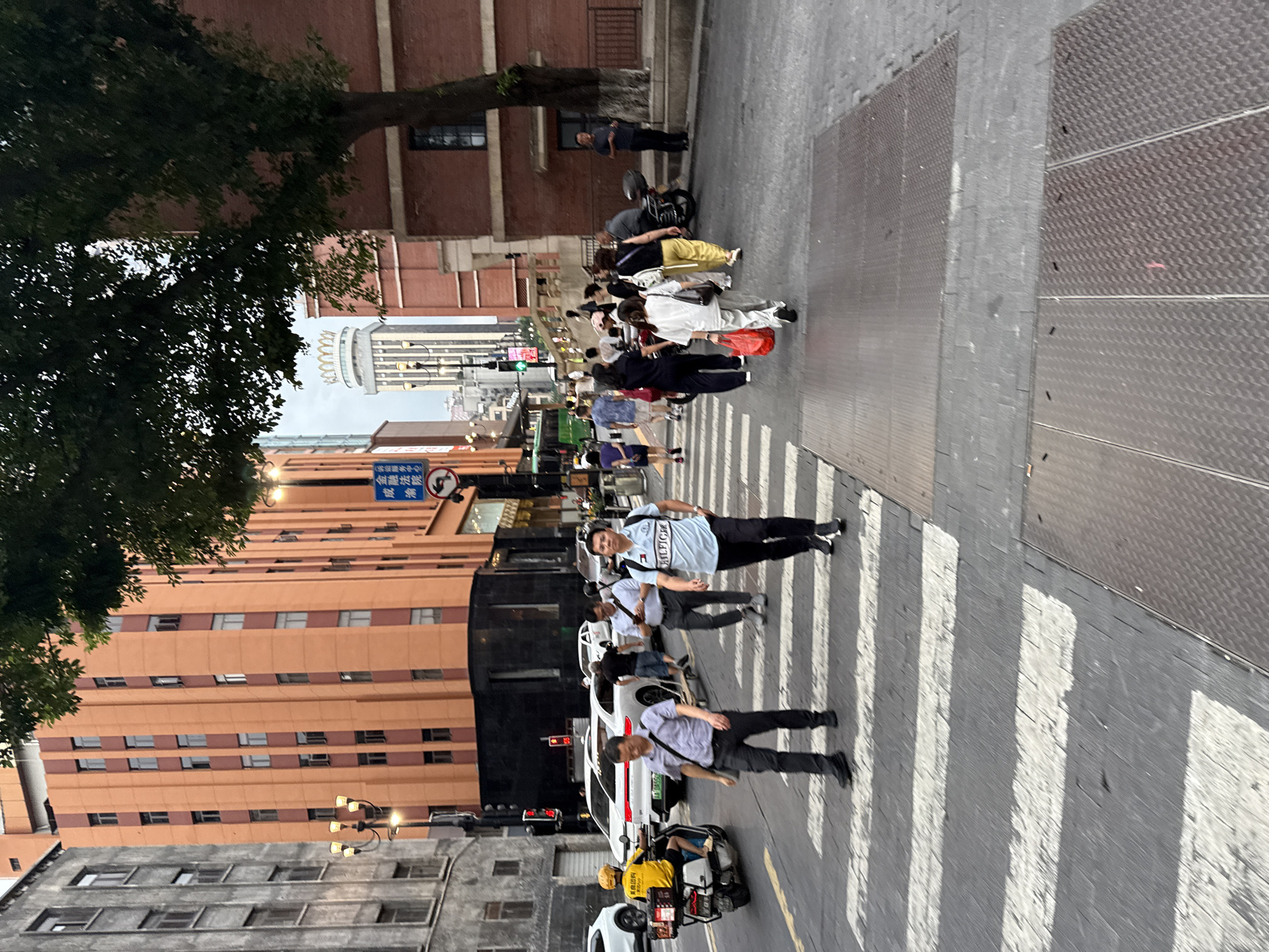 The image depicts a street scene in Yuzhong District, China, characterized by urban architecture and pedestrian activity. In the foreground, a zebra crossing is visible on a paved street. Several individuals are crossing the street, with some walking from left to right and others from right to left. A man in a light blue t-shirt with "HILFIGER" written on it is walking towards the viewer, while another man in a gray shirt with a shoulder bag is walking away. To the left, a person in a yellow uniform rides a scooter with a delivery box.
The background features multi-story buildings with distinct architectural styles. One building is painted in shades of orange and brown, with a vertical emphasis created by contrasting colored panels and numerous windows. Adjacent to it is a gray building with a more uniform facade. Further in the distance, a tall building with a distinctive crown-like structure on top stands out. The street is lined with lampposts, and traffic lights are visible, indicating an active intersection. Trees with green foliage are present, one large tree prominently positioned to the right of the frame, with its branches extending over the street. The sky is overcast.