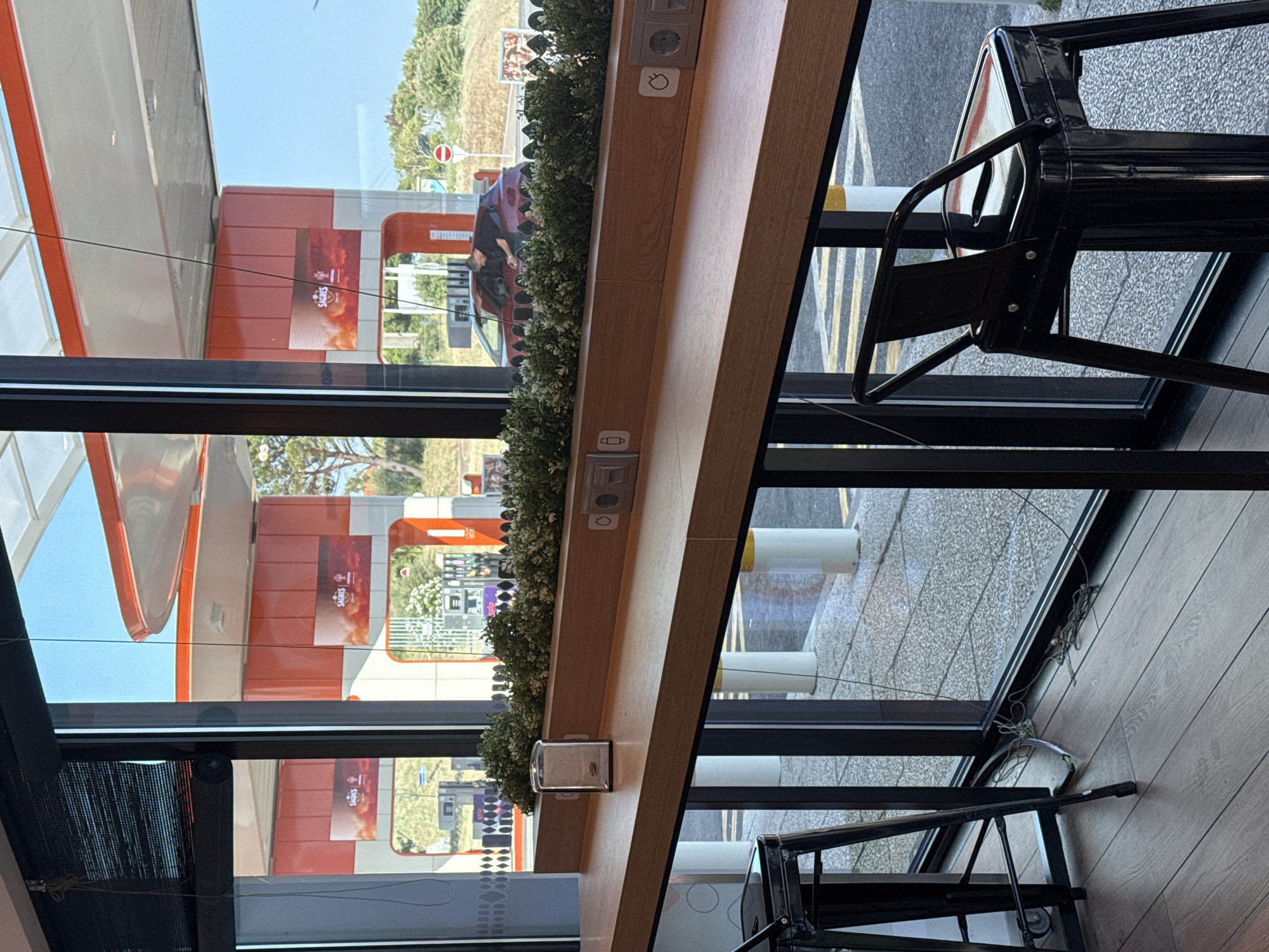 The image depicts an interior view looking out through a large window at a gas station, likely located in Oeiras, Portugal, given the prominent "Galp" branding on the gas station structures, which is a major Portuguese energy company. The window forms a long counter, made of a light-colored wood-like material, with two black metal stools placed in front of it. Mounted on the counter are electrical outlets and USB ports. A glass napkin dispenser sits on the counter near the left edge.
Outside the window, the gas station features several orange and white structures, with large banners displaying the "Galp" logo and advertisements. Gas pumps are visible, and a red car is parked at one of them, with a person leaning into the car from the passenger side. The environment appears to be sunny, with a clear blue sky. Lush green bushes are planted along the exterior of the window, forming a decorative border. In the foreground, on the left, a portion of another black metal stool is visible, and on the floor, electrical cords are tangled. The flooring inside the establishment appears to be dark wood planks.