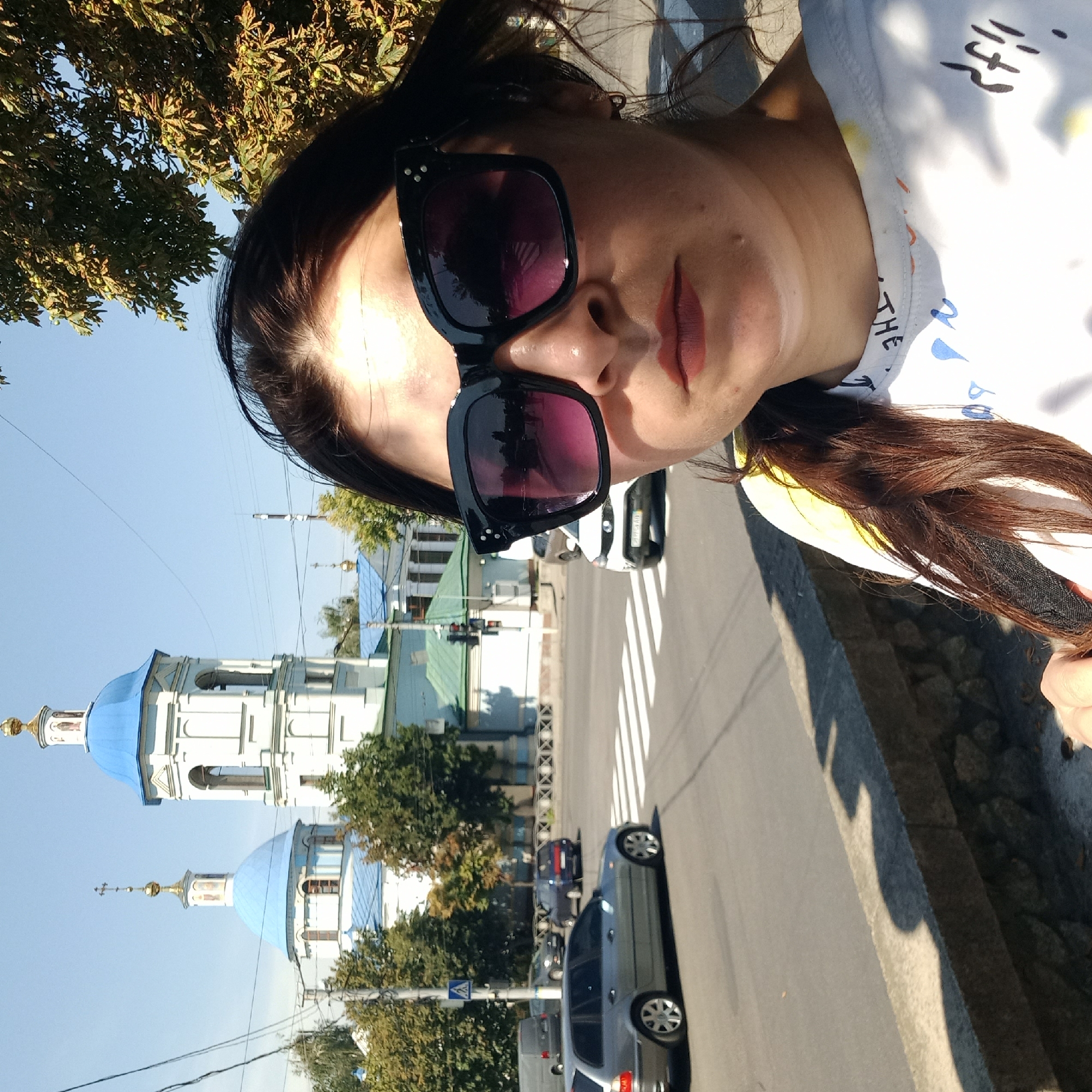A selfie is taken by a woman in Kropyvnytskyi, Ukraine, with a church in the background. The woman, wearing large sunglasses and a white t-shirt with colorful graphics, stands on a sidewalk. Behind her, a street with a pedestrian crossing is visible, with cars driving by. The prominent background feature is a white church with bright blue domes, characteristic of Orthodox Christian architecture, under a clear blue sky. Trees with green and yellowing leaves flank the church and sidewalk. The scene appears to be captured on a sunny day in a city setting.