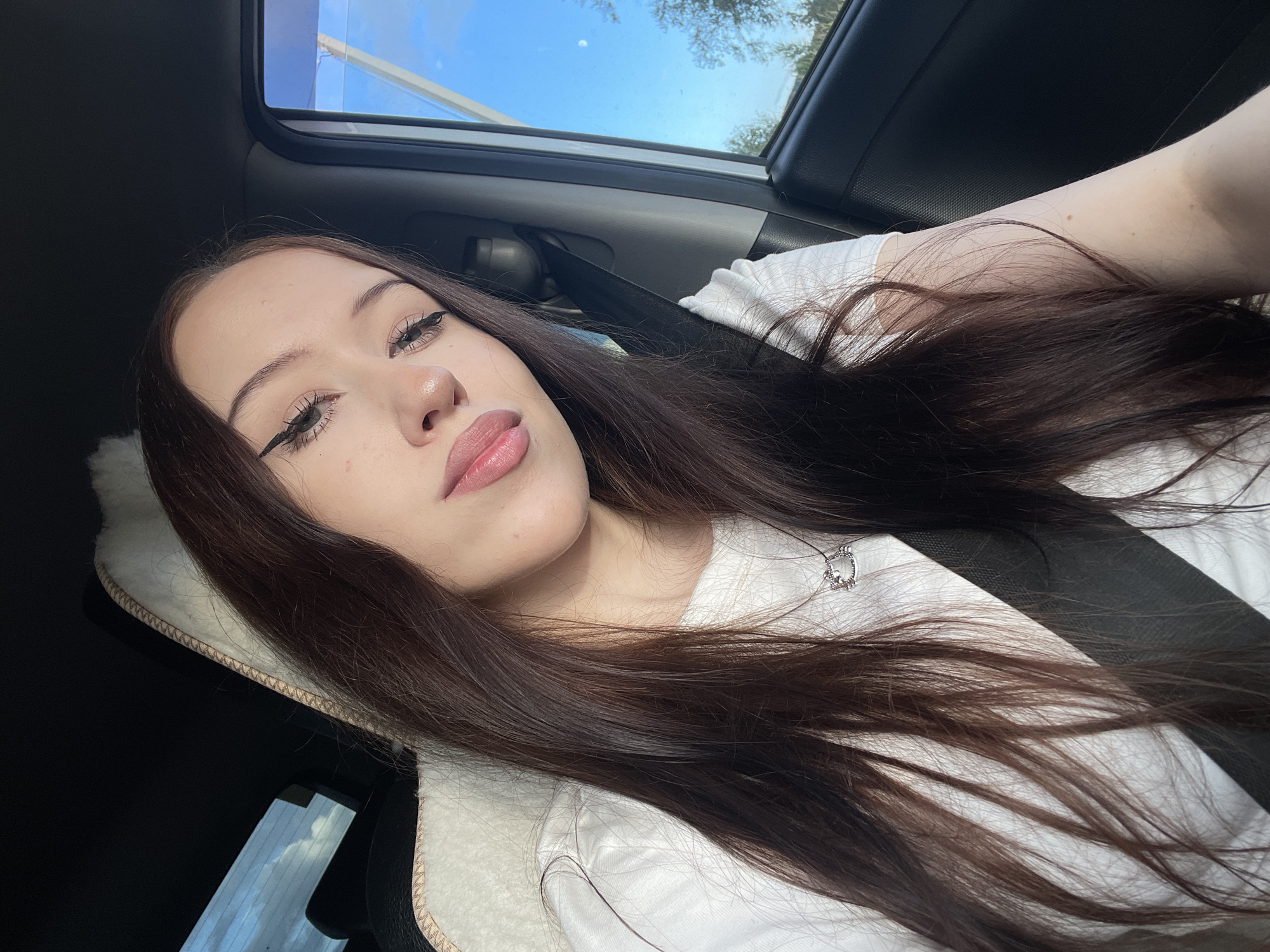 A young woman with long, dark brown hair, wearing a white t-shirt and a silver necklace, is shown from the chest up, inside a car. She is looking at the camera with a slight smile, her makeup including winged eyeliner and lip gloss. The seatbelt is visible across her chest. Outside the car window, a bright blue sky with some clouds and trees can be seen. The interior of the car is dark, with a fuzzy sheepskin-like headrest visible. The image appears to be a selfie taken during the daytime, possibly in a suburban or rural area given the natural scenery visible through the window. The specific location in Gavrilovka, Russia, cannot be determined from the visual information alone.