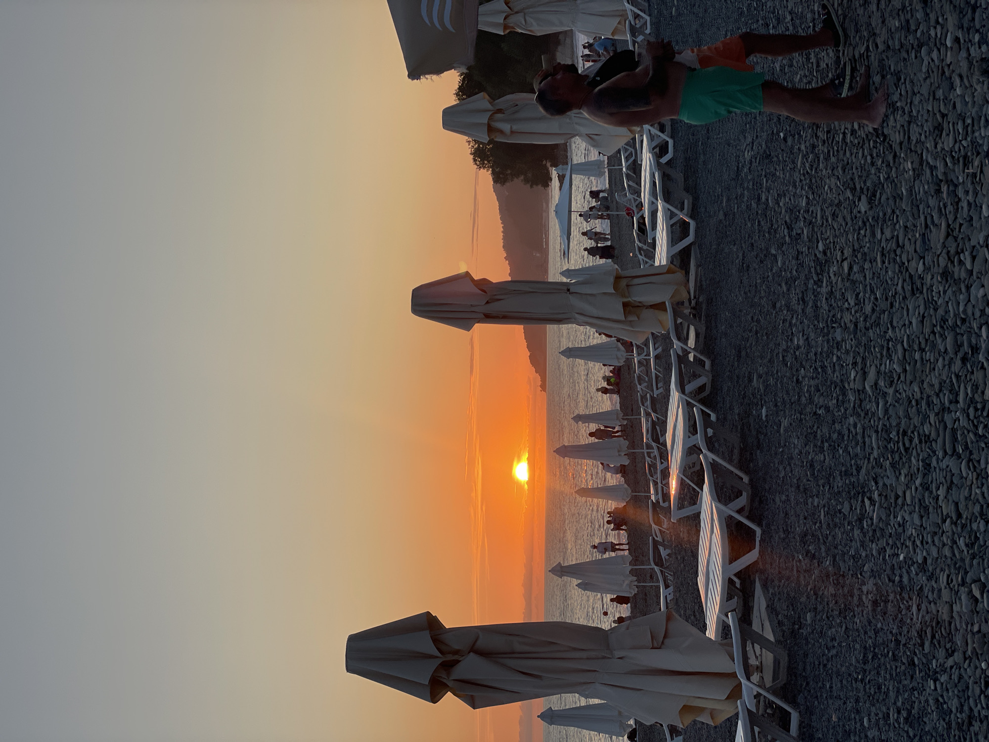 A view of a beach in Sochi, Russia, at sunset. The sun is setting over the Black Sea, casting a warm orange glow across the sky and water. There are many closed beach umbrellas and lounge chairs on the pebbly beach. Several people are visible in the background, some walking and others sitting. In the foreground, two men are standing close together, with one man with green shorts embracing the other. The scene evokes a sense of peace and relaxation.