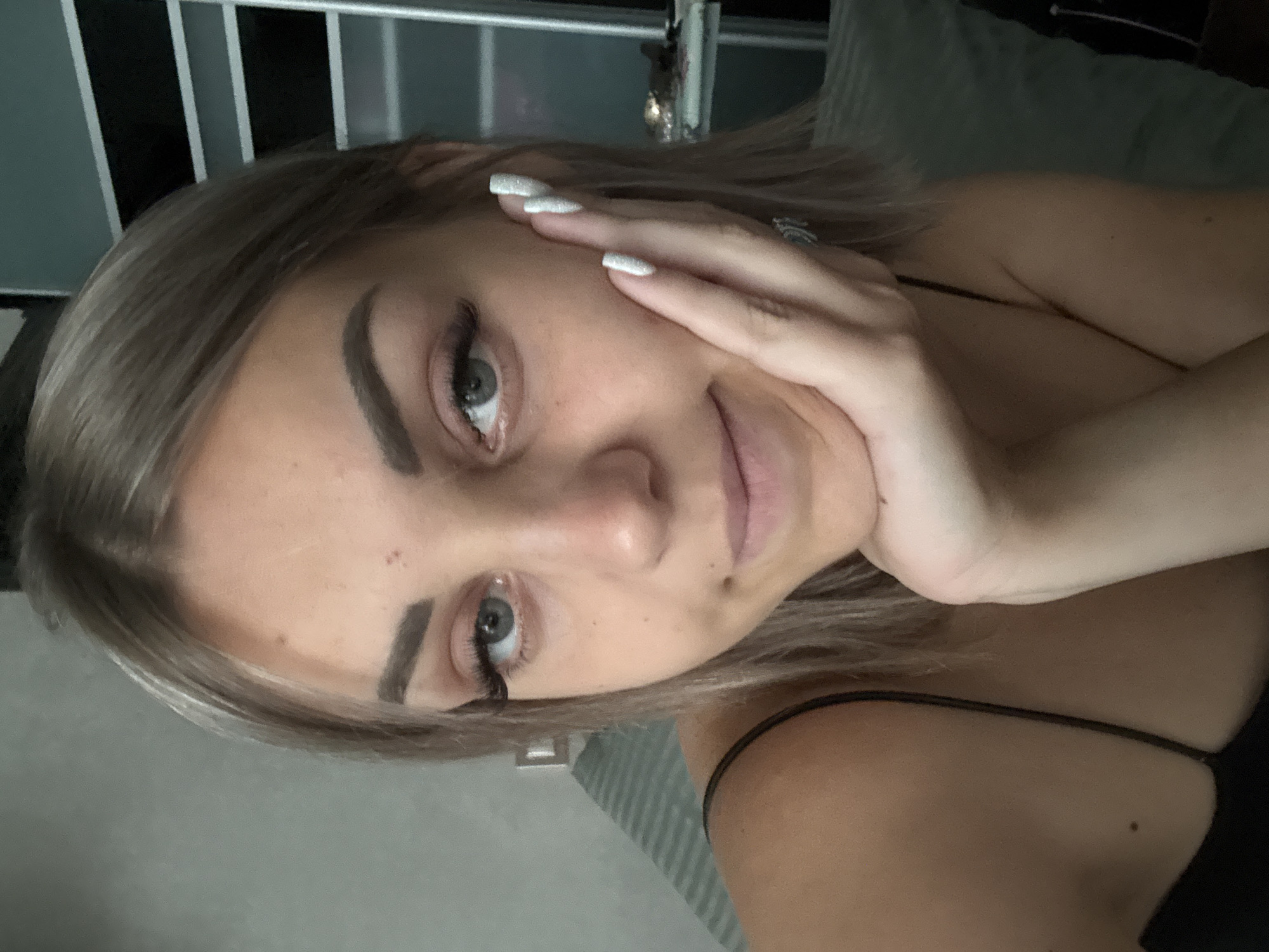 The image is a selfie of a young woman with blonde hair, blue eyes, and full lips. She is resting her chin on her hand, which has long, white, acrylic nails. She is wearing a black spaghetti strap top. In the background, there is a gray wall with what appears to be a shelf unit. The lighting is soft and natural, suggesting an indoor setting. The image does not contain any information that would allow for the identification of the location as Yuzhno-Sakhalinsk, Russia.